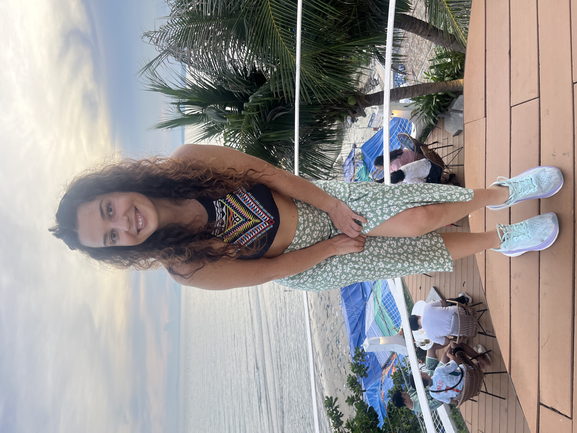 The media file captures a young woman with curly brown hair and a wide smile, posing for a photo on a wooden deck. She is wearing a black bikini top with a colorful geometric pattern and a light green skirt adorned with small white flowers. Her right foot is clad in a light blue athletic shoe with purple accents, and her left leg is bent.
Behind her, a railing separates the deck from a view of a beach and the ocean. Palm trees with lush green fronds are visible, indicating a tropical or sub-tropical location. The beach below is dotted with people and numerous blue and white tents, suggesting a lively tourist area or market. The ocean is calm, with gentle waves lapping at the shore. The sky is a soft blend of blues and grays, hinting at either early morning or late afternoon.
In the background, on the deck, other individuals are seated at small tables, some appearing to be engaged in conversation. They are seated in wicker chairs. One person wears a white t-shirt, and another a patterned shirt. The overall atmosphere appears relaxed and vacation-like. The scene is set in Mũi Né, Vietnam, a location known for its beaches and resorts.