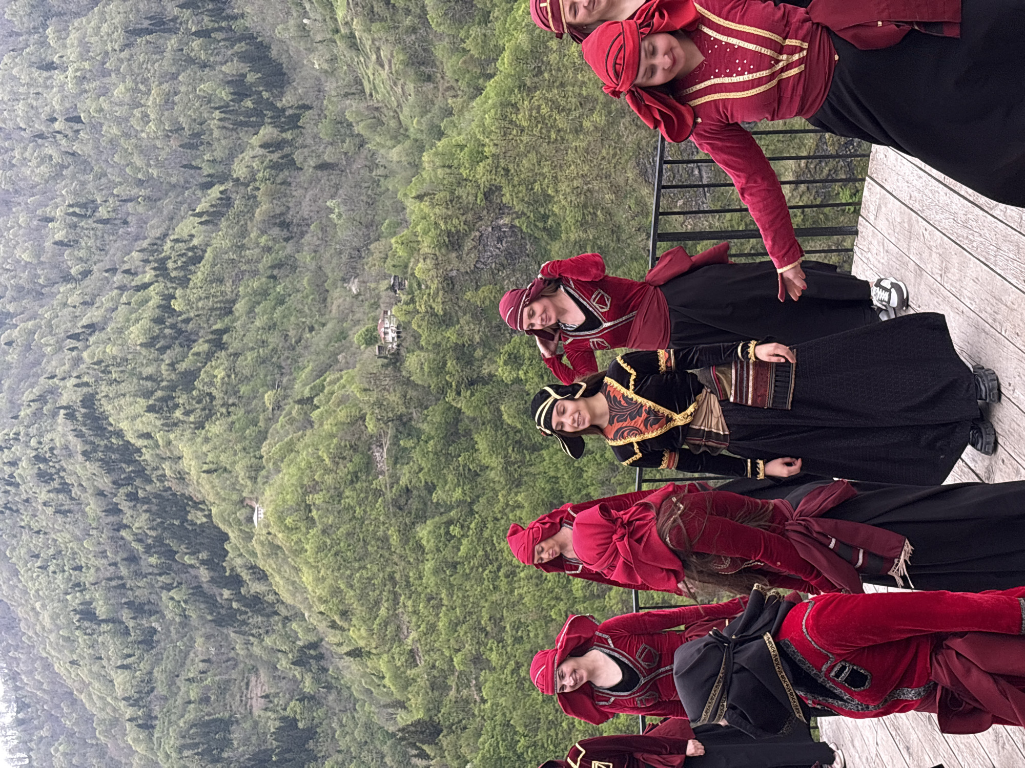 On a wooden platform overlooking a lush, mountainous landscape near Kveda Chkhutuneti, Georgia, a group of approximately six women stand in a staggered line, likely posing for a photo. All are wearing traditional Georgian attire consisting of long black dresses, and red and gold embellished velvet jackets. The women are smiling and appear relaxed. The backdrop shows a steep mountainside covered in dense green forest, dotted with small buildings. The weather appears overcast and hazy. A simple black metal railing separates the platform from the steep drop-off.