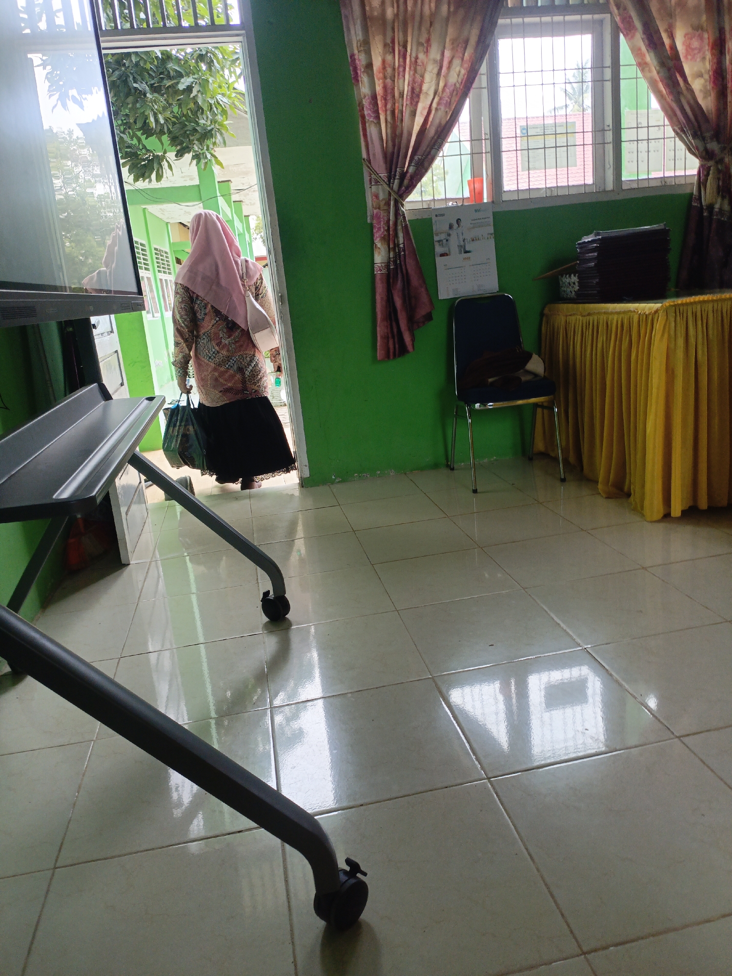 The image depicts the interior of a room, possibly a classroom or meeting space, with a woman wearing a pink hijab and a patterned shirt walking through a doorway. The room has green walls, tiled floors, and a window covered with floral curtains. In the foreground, there is a large, dark-colored mobile screen on a stand with wheels. To the right, a yellow tablecloth covers a table, and a dark blue chair with a brown item on it is placed nearby. A stack of dark items, likely folders or books, rests on the table. Outside the doorway, the exterior of a building with green pillars and windows is visible. The scene appears to be located in Lhokseumawe, Indonesia, based on the context provided.