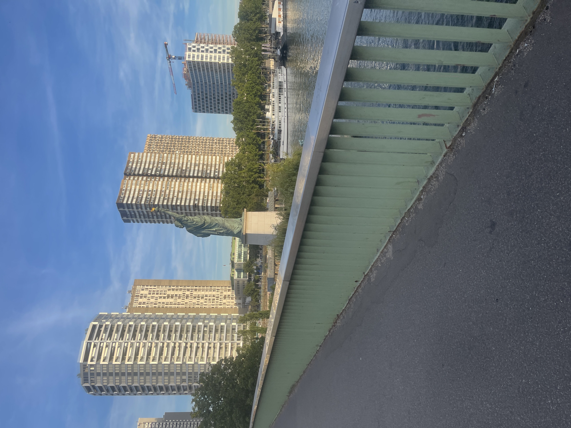 The photograph captures a cityscape in Paris, France, focusing on the Seine River and its embankment, with several modern high-rise buildings forming the backdrop. In the foreground, a paved walkway runs alongside a green metal railing that extends into the distance. The walkway appears to be part of a bridge.
Dominating the middle ground is a statue of liberty, a bronze replica, standing on a stone pedestal. Behind the statue, the Seine River flows, with several boats docked along the bank. One prominent white boat, likely a river cruise vessel, is docked closest to the camera. Trees line the riverbank, providing a touch of greenery.
The background is characterized by a cluster of tall residential buildings, exhibiting distinct architectural styles. The building on the far left is a white cylindrical tower with many balconies. Adjacent to it is a taller, beige-colored rectangular building with numerous windows. Further to the right, another similar beige building is visible. In the far right of the image, a newer, curved white building under construction is present, topped by a construction crane.
The sky is a clear blue with wispy white clouds, suggesting fair weather, and the sunlight casts soft shadows, indicating it might be late afternoon or early morning. The overall scene depicts a modern urban environment integrated with the historical element of the Seine River and its iconic waterways.