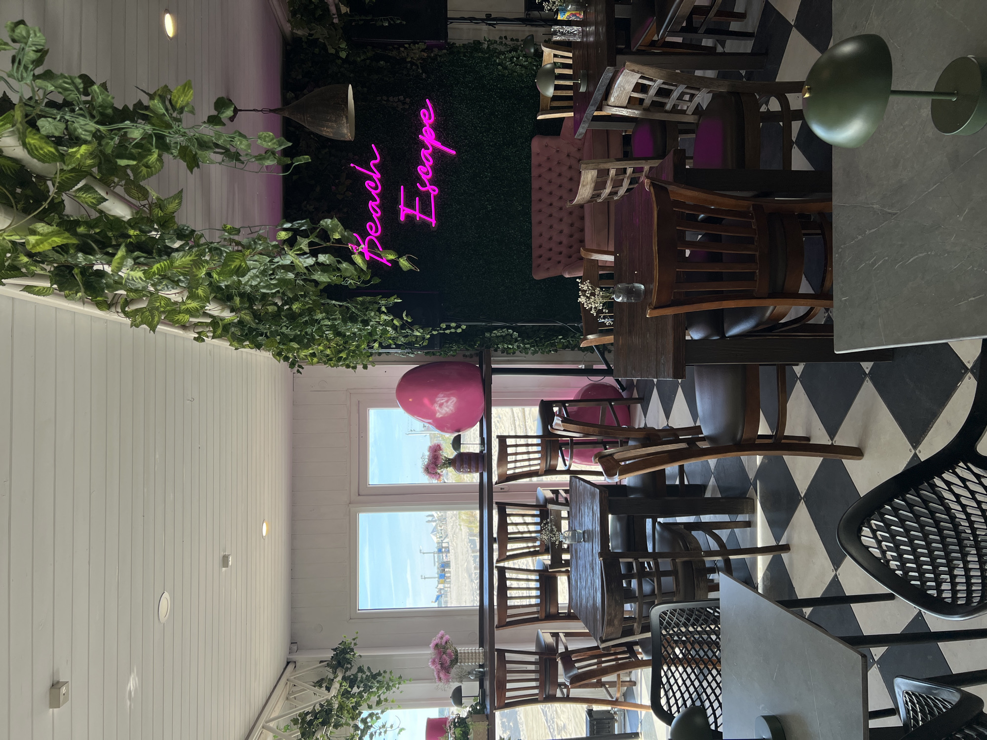 The image depicts the interior of a beachside cafe with a view of a sandy beach and the ocean through large windows. The cafe features a checkered black and white tiled floor. A long wooden bar with high stools overlooks the beach. Several wooden tables and chairs are arranged throughout the space.
A notable feature is a neon pink sign on a wall covered in artificial greenery that reads "Beach Escape". In front of this wall, there is a plush pink sofa and two wooden chairs. Large pink balloons, one in the shape of a heart, are visible. Green artificial vines hang from the ceiling.
Two small green lamps with metallic bases are placed on some of the tables. In the foreground, a dark grey table with a similar green lamp is present. The overall atmosphere is casual and evokes a beach resort vibe.
It is not possible to determine the city of Liepaja, Latvia, from the provided image.