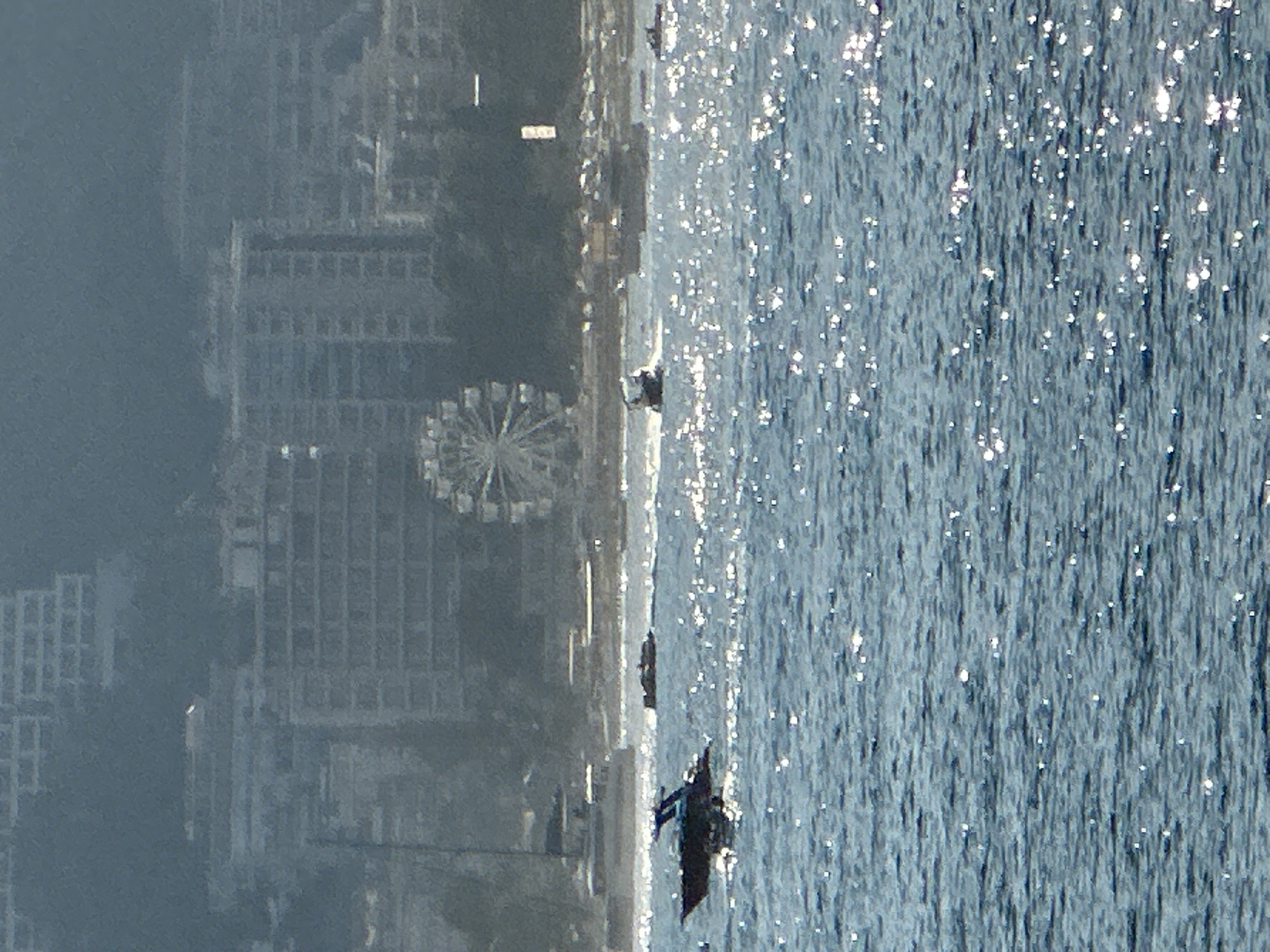 A wide shot captures a section of the Adriatic Sea with a coastline in the background. The water is choppy and reflects sunlight, creating many bright highlights. In the foreground, a dark-colored motorboat with a blue canopy is moving from left to right. Further back, other smaller boats are visible on the water. The coastline features a Ferris wheel, numerous multi-story buildings, and some vegetation. The overall atmosphere is hazy, suggesting distance or atmospheric conditions. This scene is located in Budva, Montenegro, a coastal city known for its beaches and historic old town.