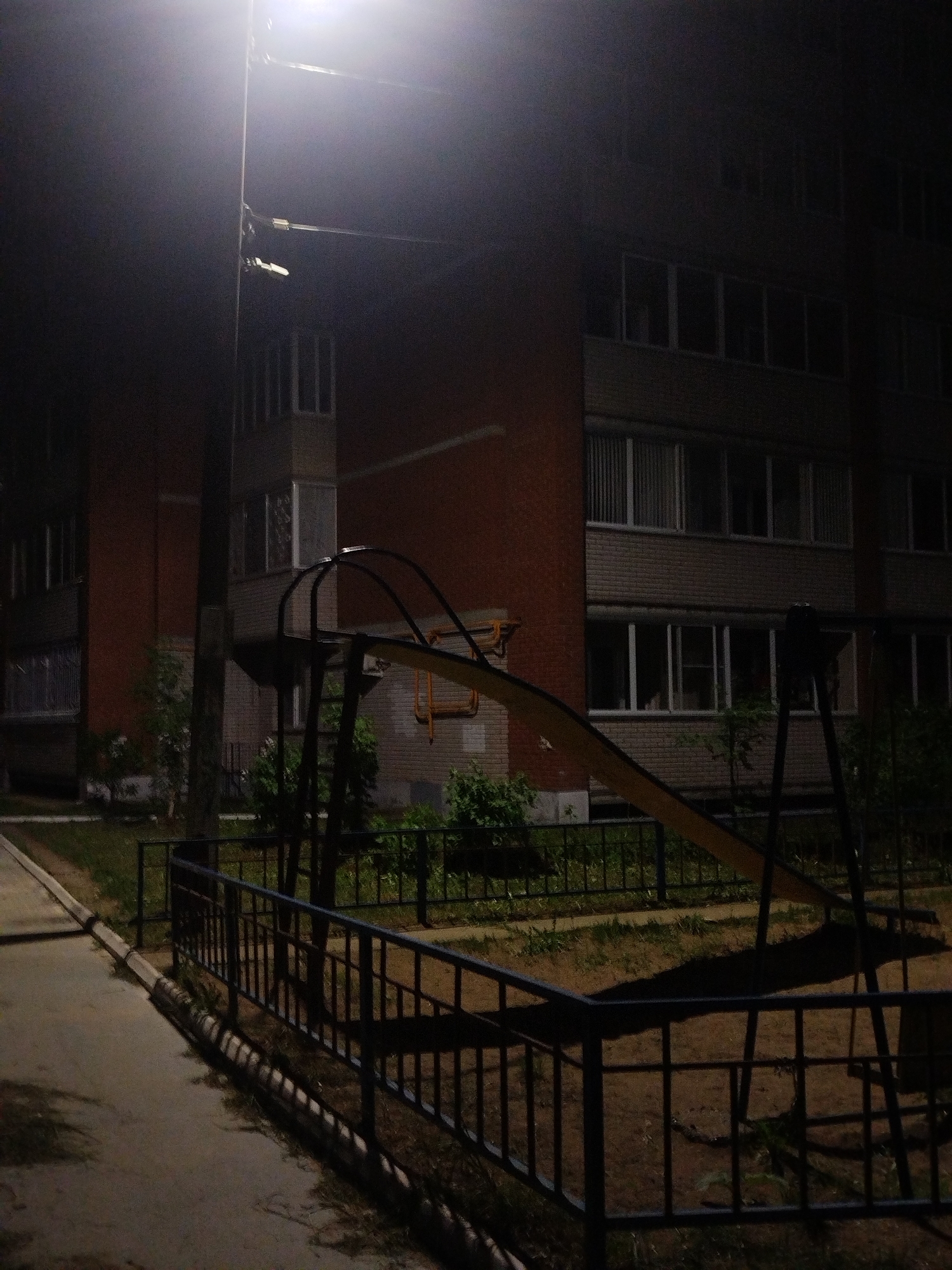 The image shows a nighttime scene in an urban environment, specifically a playground next to a multi-story brick building. The scene is illuminated by a bright street lamp positioned near the playground equipment. The playground includes a slide, visible over a low fence. The area beyond the fence features some greenery. The building has several rows of windows with blinds or curtains visible in some of them. The light from the street lamp casts strong shadows, adding contrast to the scene. The overall impression is one of a quiet, still night in a residential area.