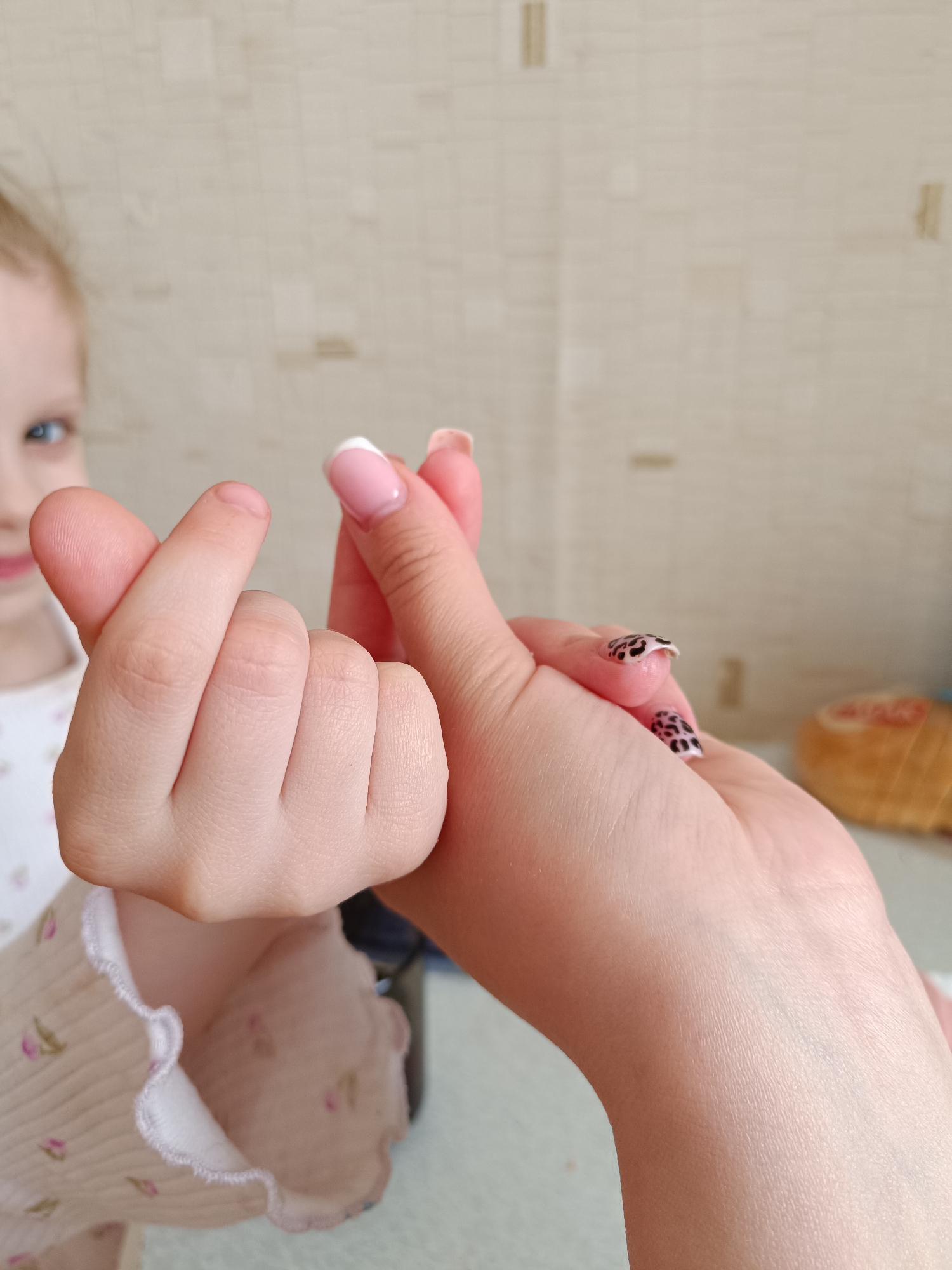 A child and adult create a "finger heart" symbol with their hands. The child's hand is on the left, while the adult's hand is on the right. The child is wearing a white shirt with pink flowers. The adult has pink nail polish with white tips on one hand and a leopard print design on the other. A blurred face of a young child is visible in the background. The picture was taken inside. No specific location within Tobolsk, Russia is discernible from the image.