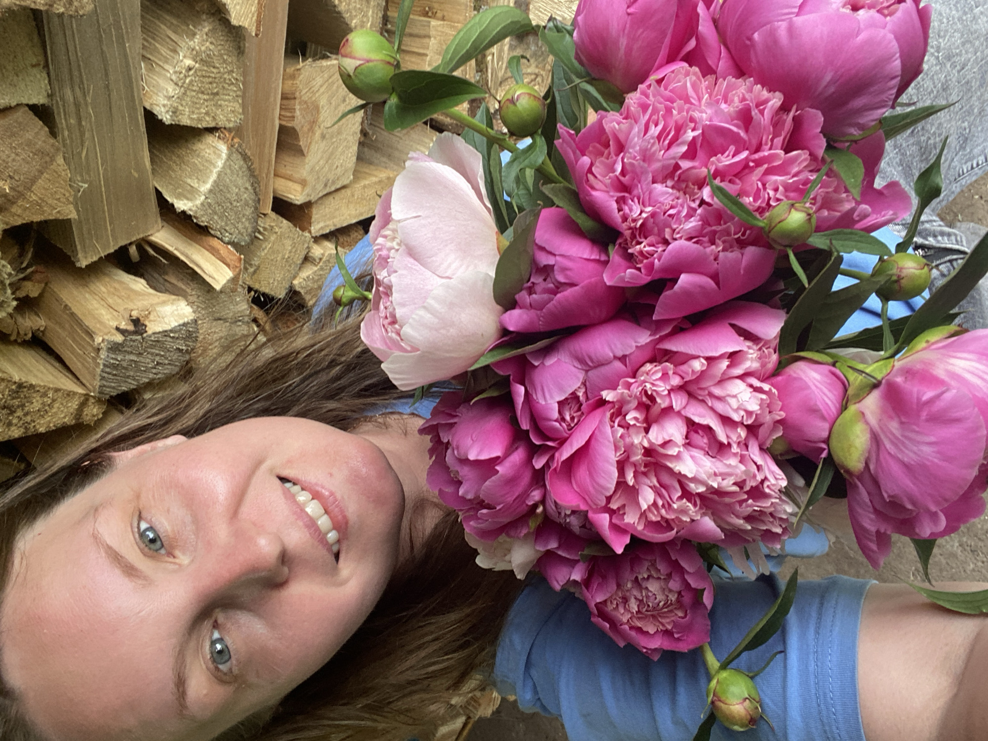 A woman with light brown hair and blue eyes, smiling, is holding a bouquet of pink peonies in front of a stack of firewood. The woman is wearing a blue shirt. The image appears to be a selfie taken outdoors. The location is not specified in the image or its metadata.