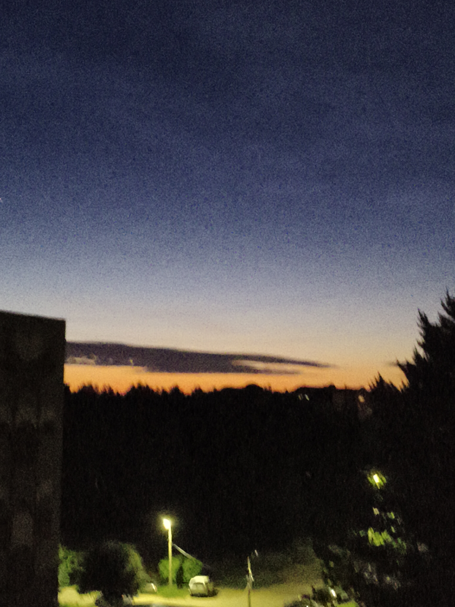 This image depicts an outdoor twilight scene in the city of Unknown, Unknown. The upper sky is a deep navy blue, gradually transitioning to a lighter blue-grey and then to a prominent horizontal band of orange and yellow light at the horizon. A dark, elongated cloud formation is silhouetted against the bright horizon, positioned centrally. The lower portion of the image is predominantly dark, featuring the silhouettes of numerous trees forming a dense line across the mid-ground. On the far left, a dark, angular structure, consistent with a building's facade, occupies the foreground. In the lower center, a bright yellow street lamp on a pole illuminates a small area, revealing green foliage and a light-colored vehicle, possibly a car or van, parked on a discernible surface. A fainter light source is visible to the right, partially obscured by trees. No human figures or specific actions are visible within the frame. The entire image exhibits digital noise, particularly noticeable in the sky and darker areas.