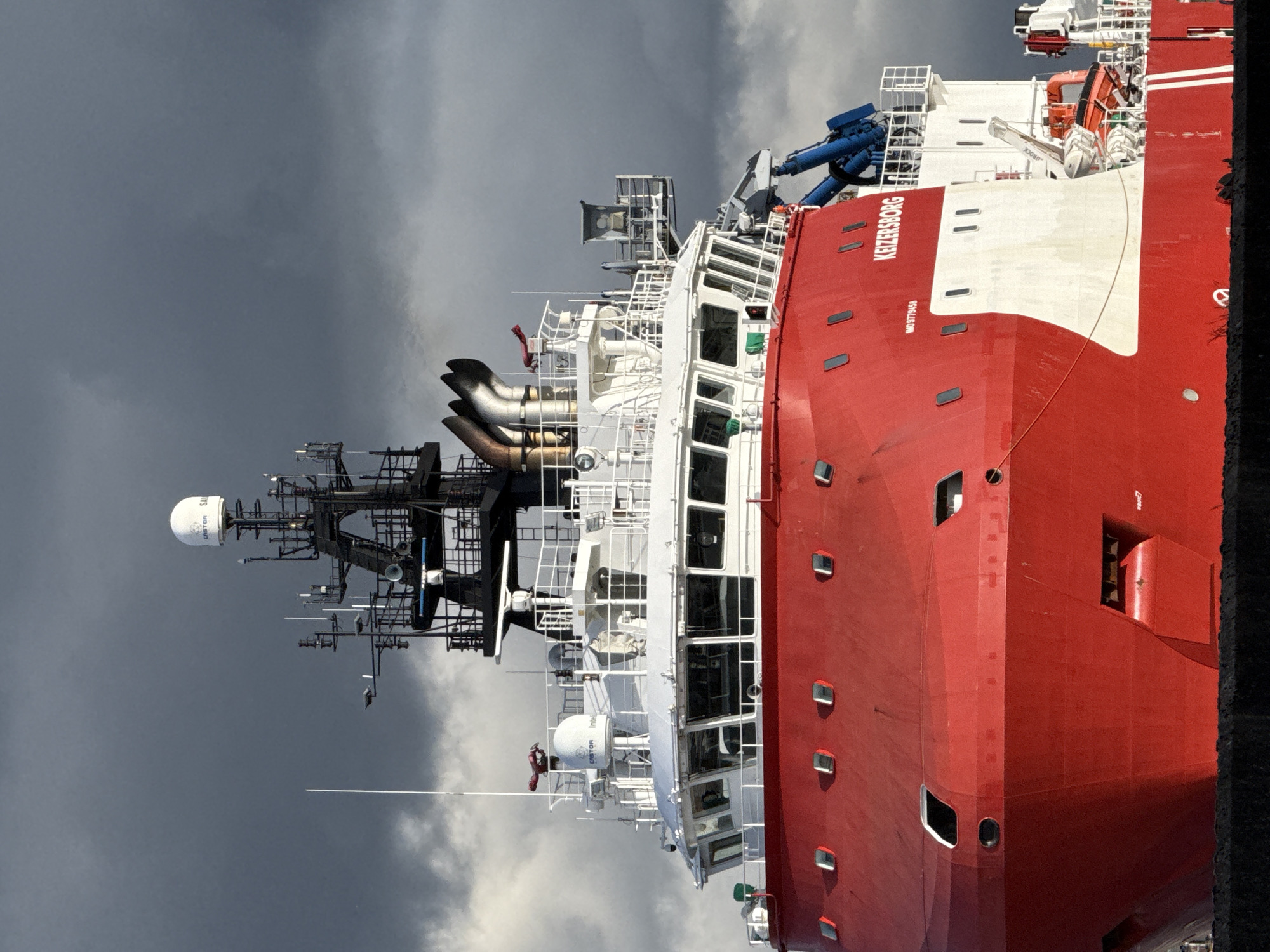 The image shows a close-up view of a red and white ship named "Keizersborg" docked in Great Yarmouth, United Kingdom. The vessel is primarily red, with a white stripe running along its length, and several small, rectangular windows. The ship's navigation and communication equipment is visible, including radar domes, antennas, and exhaust pipes. The sky behind the ship is filled with heavy, dark clouds. The hull of the ship is visible with windows. "Keizersborg" is painted in white on a red background. A black, possibly concrete, structure is visible at the right edge of the image.