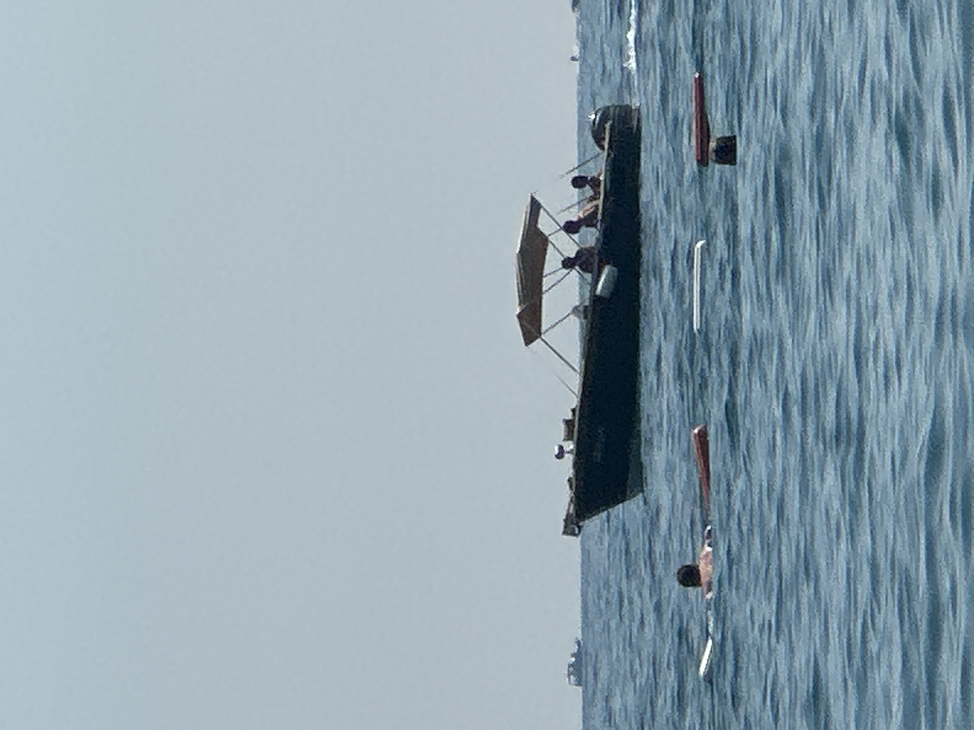 An eye-level, wide shot captures a serene scene on the water near Boreti, Montenegro. The clear, pale blue sky meets the calm, blue water, creating a vast expanse. In the foreground, a lone swimmer is partially submerged, supported by a white float. To their right, a cylindrical red object floats on the surface. Further into the frame, a dark-hulled motorboat with a tan canopy navigates the water, carrying three silhouetted figures. The boat appears to be moving away from the viewer, leaving a faint wake. In the distance, a much smaller boat is visible on the horizon. The overall impression is one of a peaceful day at sea.