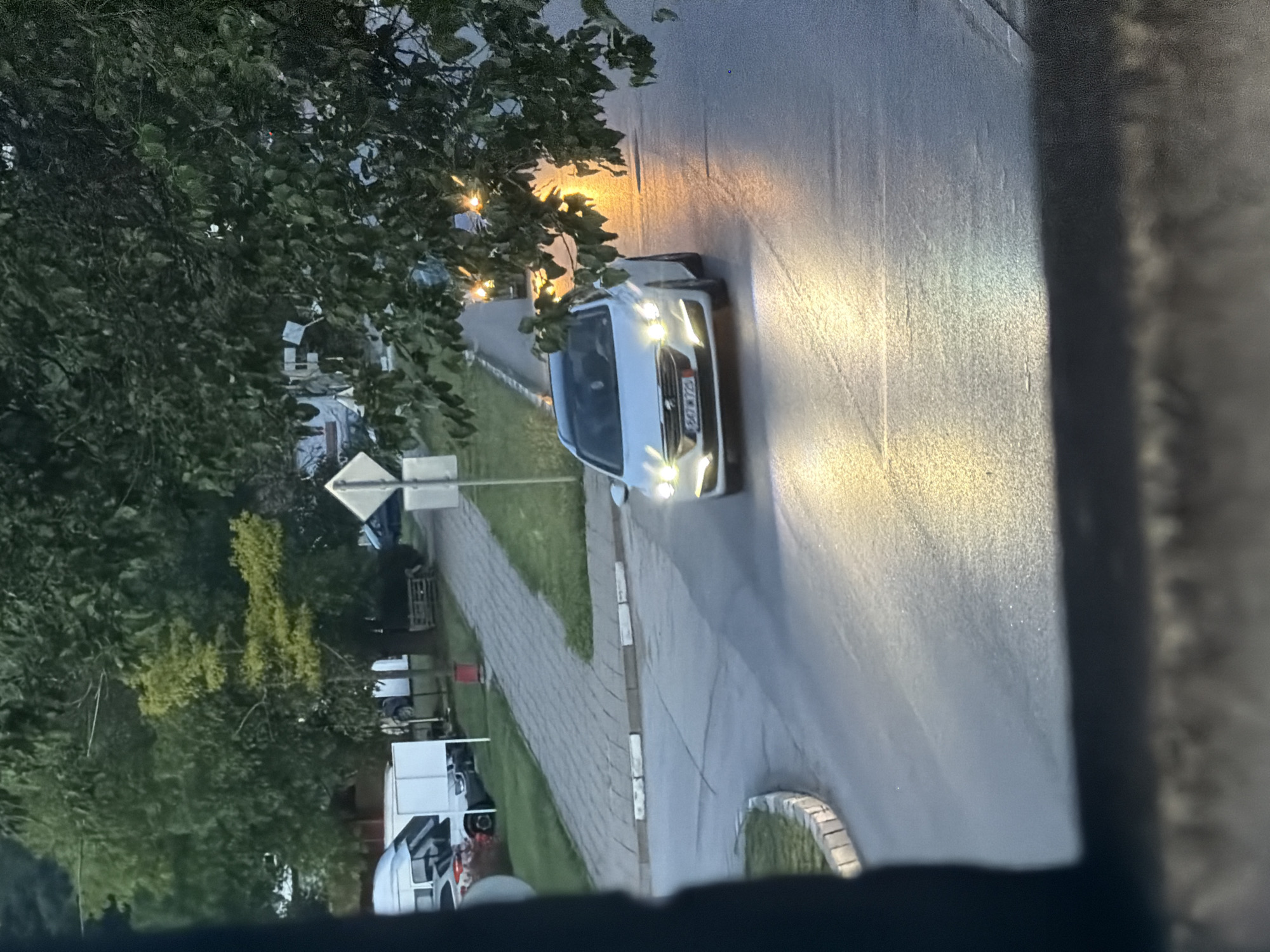 This photograph, taken in Iskar, Bulgaria, captures a white car driving on a wet, paved road at dusk or night. The car's bright headlights strongly illuminate the asphalt ahead, casting distinct cones of light. The image is framed by a dark, out-of-focus foreground element, suggesting it was captured from inside a building or vehicle.
To the left of the road, a patterned grey paver sidewalk runs parallel, bordered by a concrete curb. Dense green foliage, including trees and bushes, lines the street, with some distant buildings and other parked vehicles partially visible through the leaves. A square road sign on a pole is also present among the trees near the sidewalk. The white car's license plate is clearly visible, reading "B0 8272 KA," which identifies it as a Bulgarian registration from the Sofia region. The overall dim lighting, active headlights, and reflective road surface indicate an evening or night scene where artificial illumination is prominent. No people are visible.