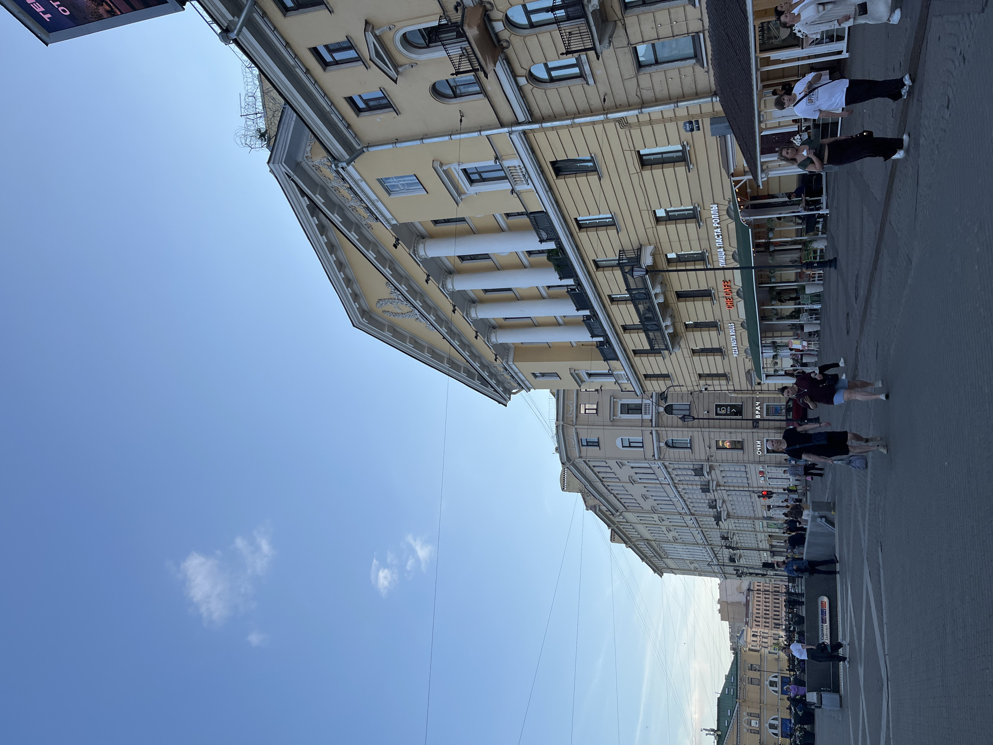 This media file shows a street scene in Saint Petersburg, Russia. The buildings lining the street are old, with light yellow facades and classical architectural elements like columns and ornate trim. The perspective is tilted, giving a sense of looking up at the buildings or at an angle from across the street.
Several people are walking on the sidewalk and street. They appear to be casually dressed and are spread out, suggesting it is a normal day rather than a specific event. The sky is blue with a few scattered white clouds, indicating fair weather during the daytime.
Visible text includes signs in Russian. One sign reads "PIZZA PASTA ROLLS" and "CHE CAFE" with Russian text "ПИЦЦА ПАСТА РОЛЛЫ" and "ЧЕ КАФЕ" which translates to "CHE CAFE". Another sign says "Очки" meaning "Glasses" and "ВРАЧ" meaning "Doctor". There is also a sign that seems to say "Sadovaya 35" indicating the street address.
The overall impression is a bustling urban environment with historic architecture and people going about their daily activities.
