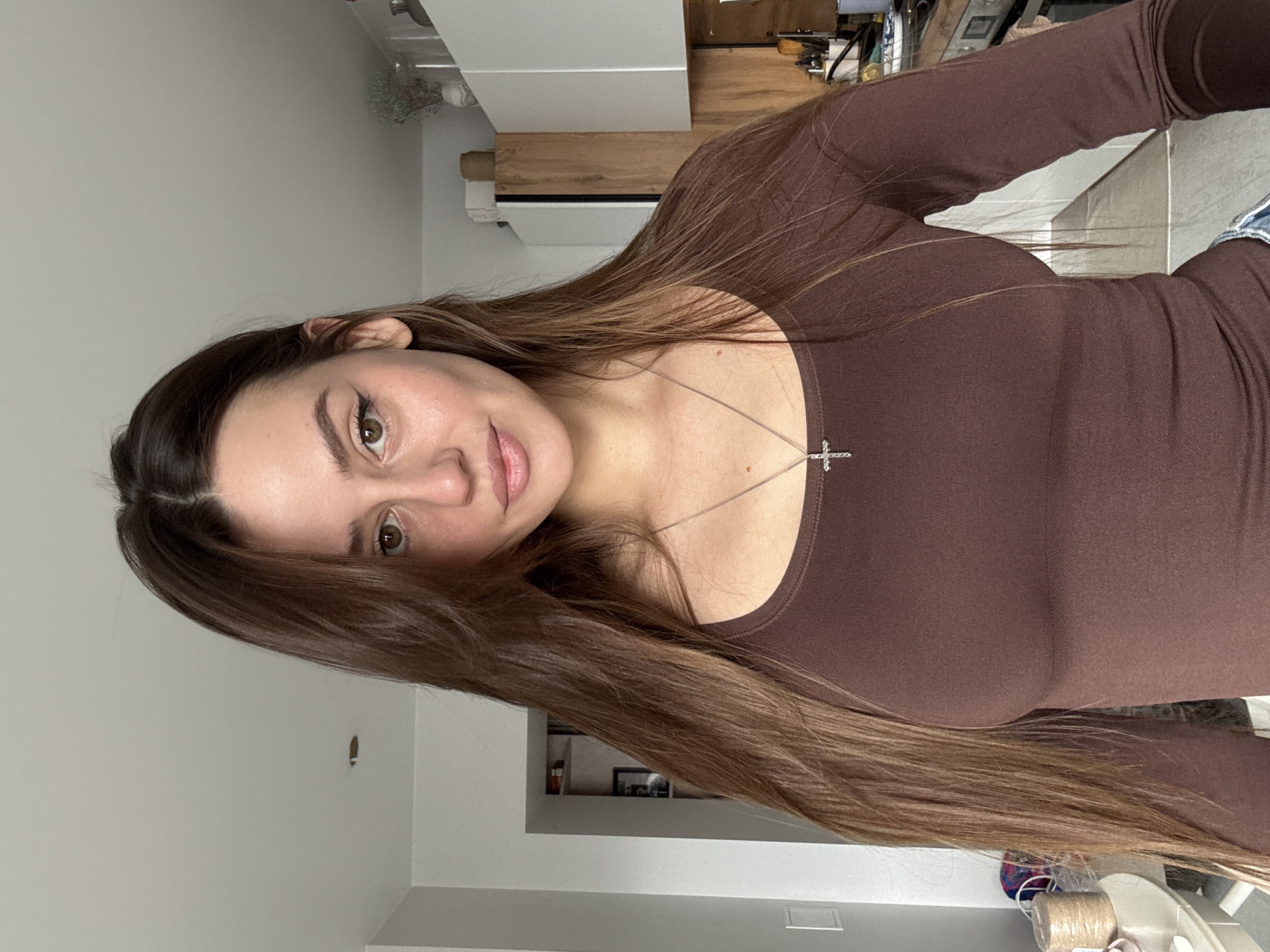 The image shows a young woman with long, brown hair, wearing a brown long-sleeved top and a silver necklace with a cross pendant. She is indoors, likely in a residential setting, as suggested by the presence of a kitchen area with cabinets, appliances, and various items on the countertop in the background. The lighting is soft, indicating either natural light or diffused artificial lighting. The exact location within the city of Krasnoyarsk, Russia is not possible to determine from the image.