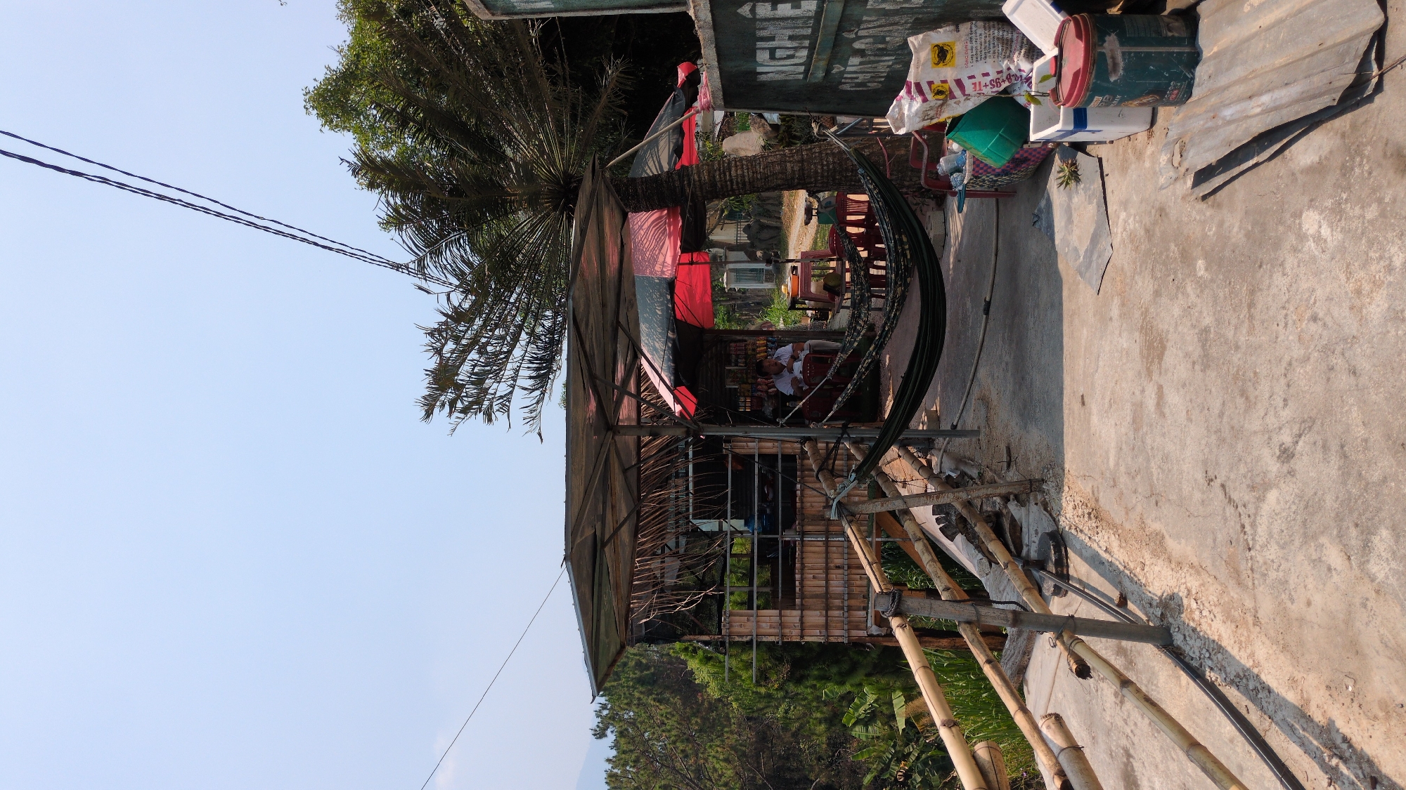The image shows an outdoor area in Lâm Sơn, Vietnam. A small, makeshift structure with a roof and bamboo supports serves as a gathering place. Inside, several people sit at tables and chairs. A hammock is strung up nearby. The area is adjacent to a concrete walkway with scattered objects like a paint can and bags. A palm tree provides shade over the structure. Power lines are visible in the sky. A hill with trees is in the background.