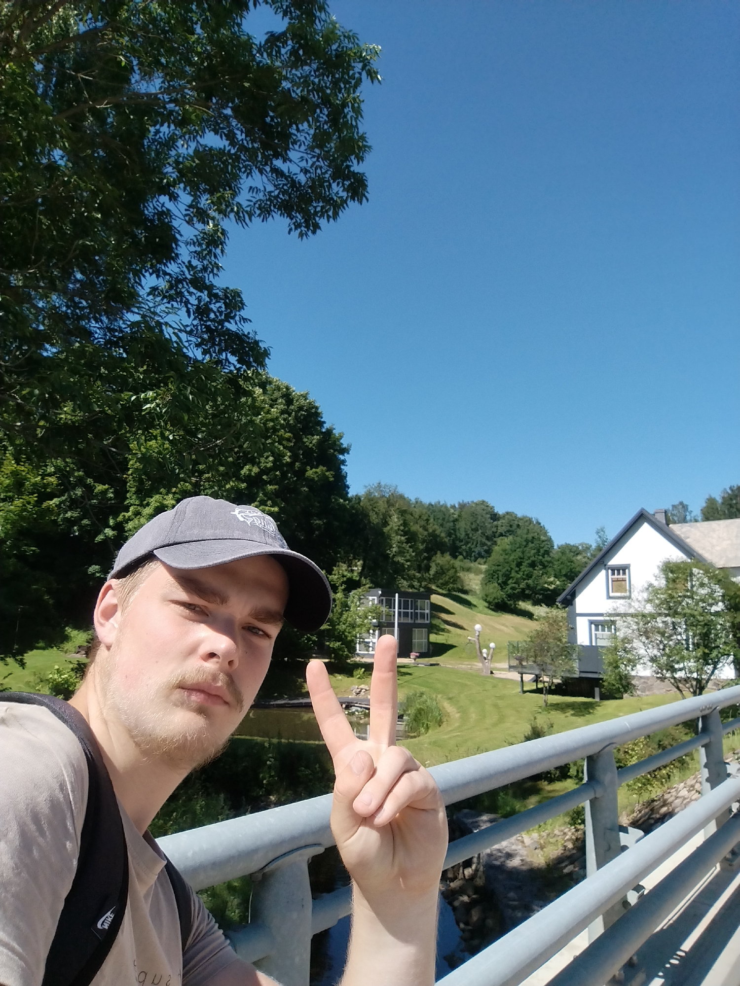 The image is a selfie taken by a young man wearing a gray baseball cap and a light-colored t-shirt, with a backpack on. He is standing on a bridge with a metal railing. His right hand is raised, forming a peace sign. The background shows a body of water, a grassy slope with trees, and two buildings. The sky is clear and blue. The context provided indicates the location is the city of Rauna, Latvia, specifically at the Rauna Mill complex.