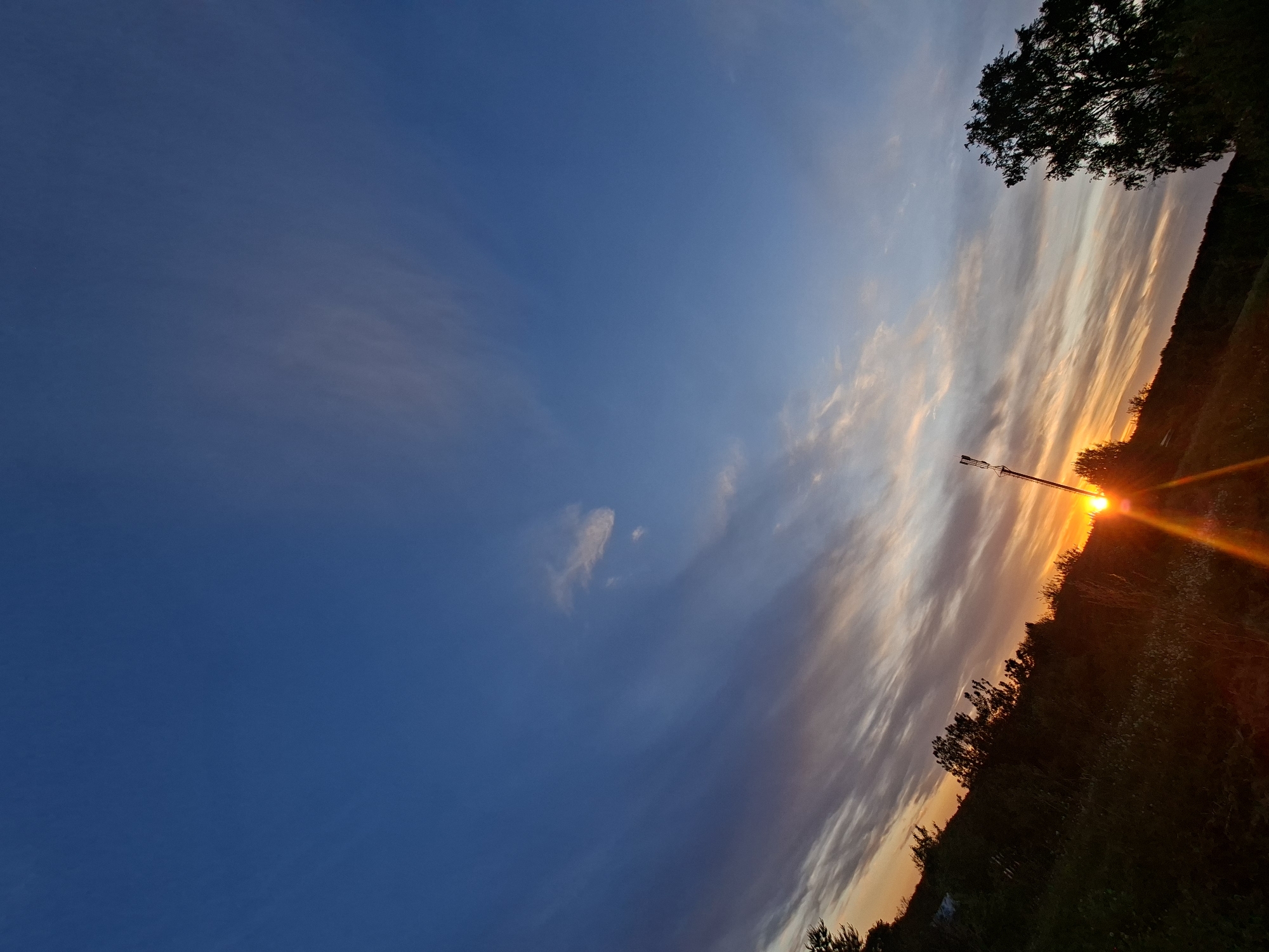 The image captures a sunset in Unknown, Unknown. The sky transitions from deep blue to orange-tinted clouds near the horizon. Silhouetted trees and a communication tower are visible on the right. The sun is positioned low, emitting bright flares. The foreground consists of a grassy area.