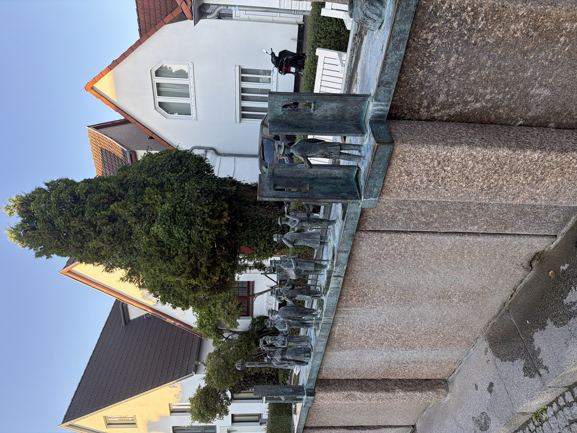 The image depicts an outdoor scene in Rostock, Germany, specifically at the "Zeitfigur" sculpture located on the Alter Strom. The sculpture, cast in bronze, features a line of figures, appearing to represent historical individuals, dressed in period clothing. The figures are arranged along a ledge that extends from a stone wall. The wall itself is constructed from large, rough-hewn granite blocks. In the background, modern buildings with white facades and red-tiled roofs are visible, along with a tall evergreen tree. The sky is clear and blue. A black scooter is parked near one of the buildings, and a dark-colored car is visible behind the sculpture. The ground in the foreground is paved with cobblestones and has wet patches, suggesting recent rain. The lighting indicates it is either early morning or late afternoon, with a warm glow on the building facades.