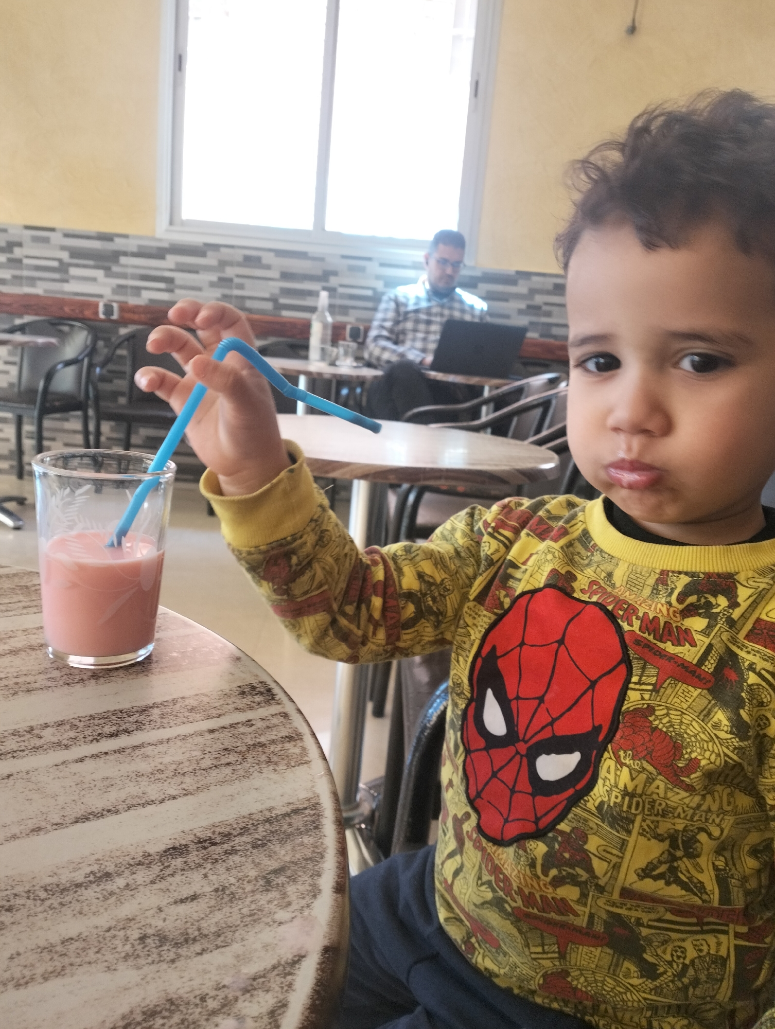 A young child sits at a table in a café in Ouaouizert, Morocco, wearing a yellow Spider-Man shirt. The child holds a blue straw above a glass of pink liquid. A man sits at a table in the background using a laptop. The cafe has white and gray tiled walls and yellow upper walls. The child looks toward the camera with a slight pout. The table in front of the child is round and has a faux wood grain top. The cafe appears to be indoors, as there is a window letting in natural light.
