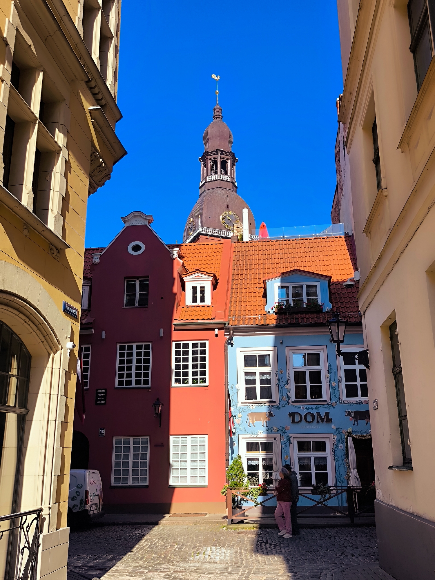 The image depicts a narrow cobblestone street in the Old Town of Riga, Latvia, with a bright blue sky overhead. The street is flanked by tall, narrow buildings of varying colors. On the left, a pale yellow building with large arched windows dominates the foreground. To its right, a narrow alley opens up to reveal two colorful buildings. The building in the middle is painted a deep red and features several white-paned windows. To its right, a vibrant blue building with white window frames and painted decorative elements, including cows and floral motifs, is visible. A sign above the entrance reads "DOM RESTORANS".
Further in the background, the dome of the Riga Cathedral, a prominent landmark, can be seen against the blue sky, with a clock face visible on its tower. The red-tiled roofs of the buildings are illuminated by sunlight, casting shadows on the cobblestone street.
Two people are standing near the blue building. One person, wearing a dark top and pink pants, is looking to the right, while the other, wearing a dark top and grey pants, is looking to the left. They appear to be conversing. A white van with a logo is parked in the alley to the left of the red building. A black vintage-style street lamp is attached to the blue building. The overall scene conveys a charming and historic urban atmosphere.