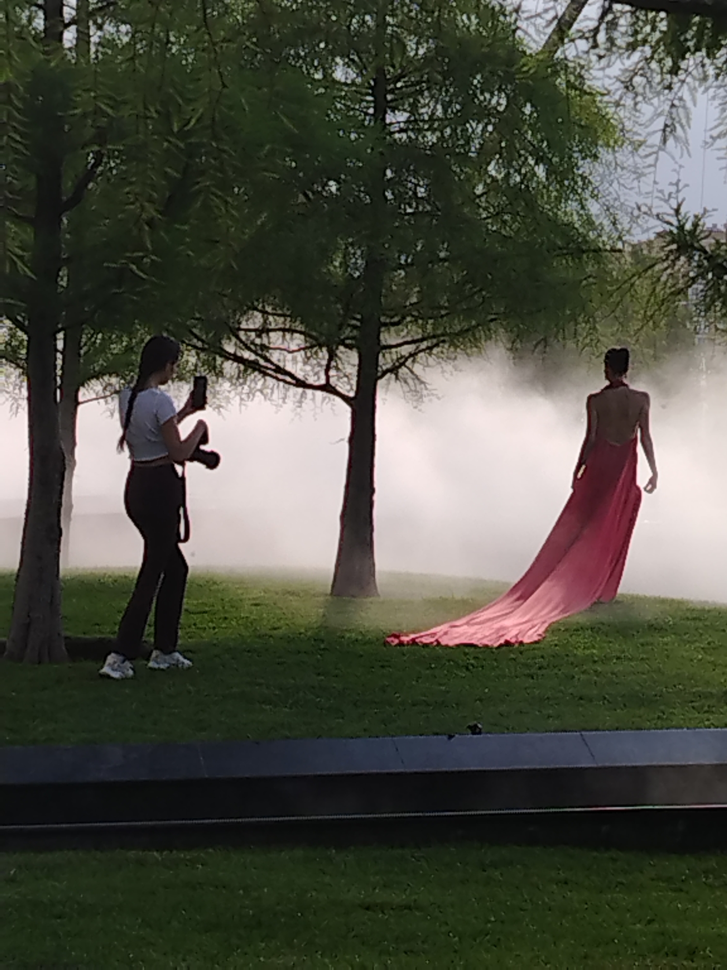 The image depicts an outdoor photoshoot with two subjects and a misty atmosphere. On the left, a woman with dark hair tied in a braid is standing, holding a camera in her right hand and a smartphone in her left. She is wearing a light-colored crop top and dark pants. She is looking towards the right, presumably at the other subject. To the right, a person, likely a woman, is posing in a long, flowing pink dress with a train that extends across the grassy ground. The person is facing away from the camera, with their back exposed. The environment is a grassy area with several trees casting shadows and a low, dark structure in the foreground. The background is obscured by a dense white mist, creating a dramatic and ethereal effect. The location is not specified as being in Krasnodar, Russia, nor is there any information in the image to suggest this location.