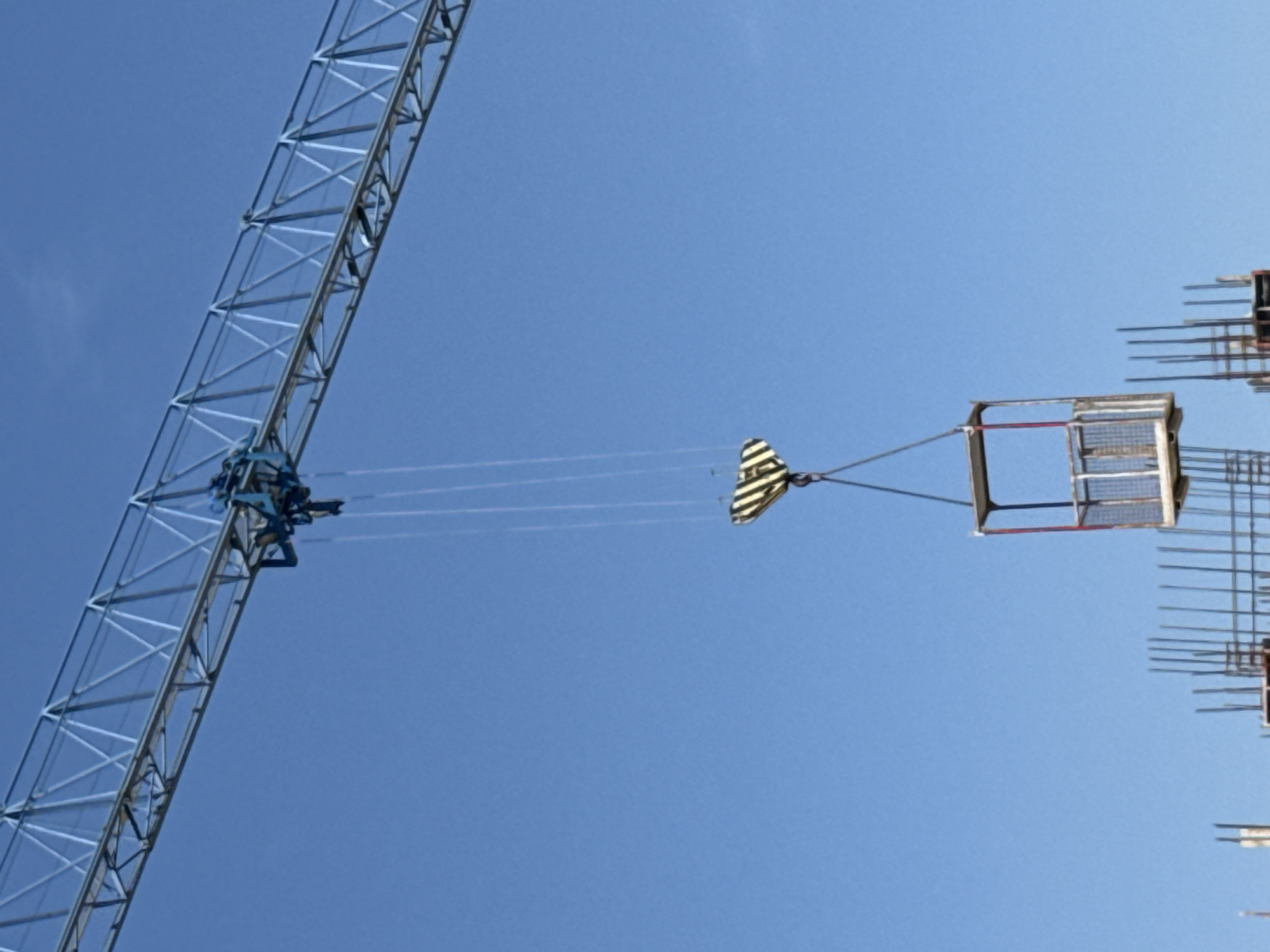 The image presents a clear, bright blue sky overhead a construction site in Varna, Bulgaria. Dominating the left side of the frame is the diagonally ascending, lattice-structured jib of a large tower crane. From its cables, a hoist block, marked with distinctive black and yellow safety stripes, is suspended. This hoist block, in turn, holds a grey, metal cage-like structure, suggesting the crane is actively involved in lifting or positioning materials for construction.
To the right, partially visible against the sky, are elements of a building under construction, characterized by numerous exposed rebar rods projecting upwards. The photograph is taken from a low-angle perspective, looking up towards the sky and the construction equipment. The weather is bright and clear, indicating daytime. No individuals are visible, focusing the scene on the industrial machinery and the nascent structure. There is no discernible text apart from the safety stripes on the hoist block.