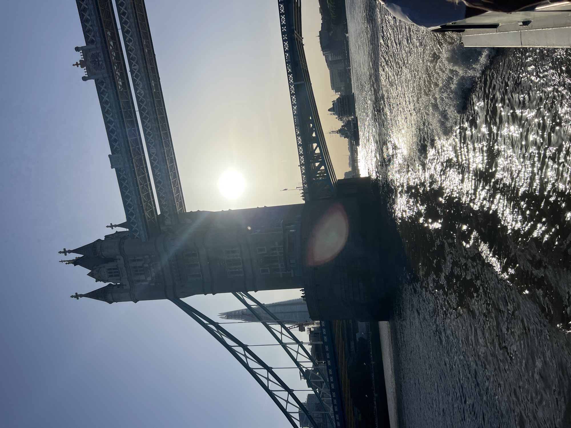 The image shows the Tower Bridge in London, United Kingdom, taken from a low angle on the water. The bridge towers are visible on the left, with the upper sections of the bridge extending towards the center. The sun is positioned behind the bridge, causing a lens flare. The Shard skyscraper can be seen through the bridge structure. The water in the foreground is sparkling due to the sunlight, and the wake of a boat is visible on the right. Some buildings can be seen on the horizon. The sky is clear and bright.