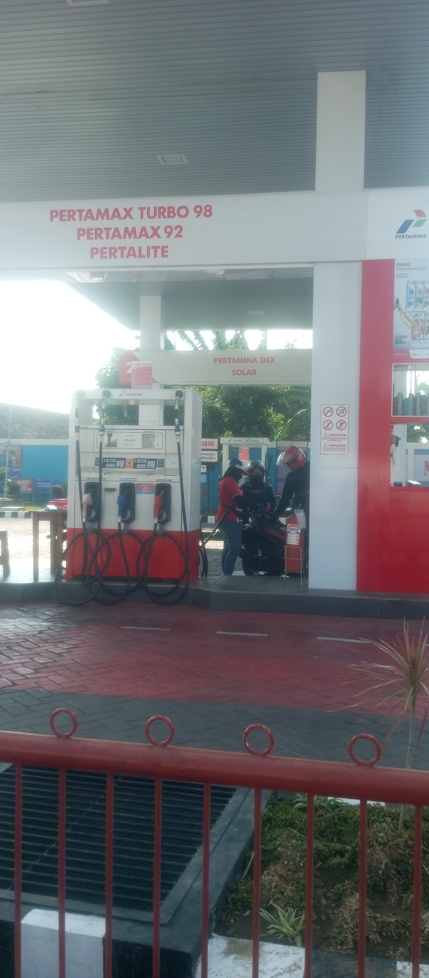 The image depicts a Pertamina gas station in Kediri City, Indonesia, under a bright, overcast sky. A white canopy with red lettering listing "PERTAMAX TURBO 98," "PERTAMAX 92," and "PERTALITE" is prominent. Below this, the Pertamina logo is visible on the right. The central focus is a gas pump island with three dispensers, each featuring red hoses and nozzles. The island has digital displays. Two people, one in a red shirt and another in dark clothing, are interacting with a motorcycle next to the pump. The ground is paved with red and gray tiles, and a red metal railing with circular decorative elements is in the foreground. In the background, greenery and a blue structure are visible.