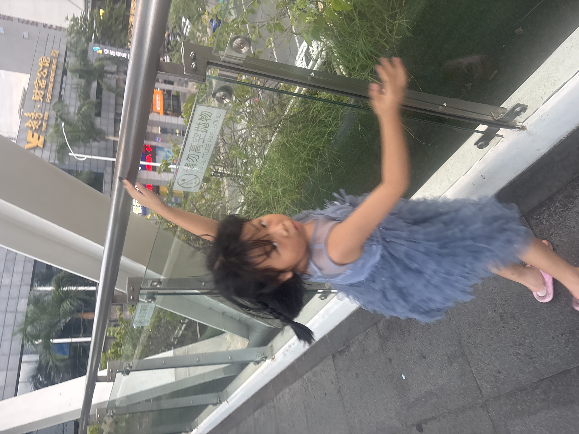 A young girl with black hair in a braid, wearing a light blue fluffy dress and pink sandals, is captured in motion on an elevated walkway. Her body is turned, with her right arm extended outwards and her left arm bent at the elbow. She is looking towards the camera with an open mouth and wide eyes. The walkway features a metal railing and glass panels, with lush green plants visible below and in the background. Behind the railing, a building with a large yellow "Y" logo and Chinese characters indicating "Fortune Apartment" is visible, suggesting the location is in the Liwan District of China. The overall scene depicts a candid moment of a child engaged in playful movement on an urban overpass.