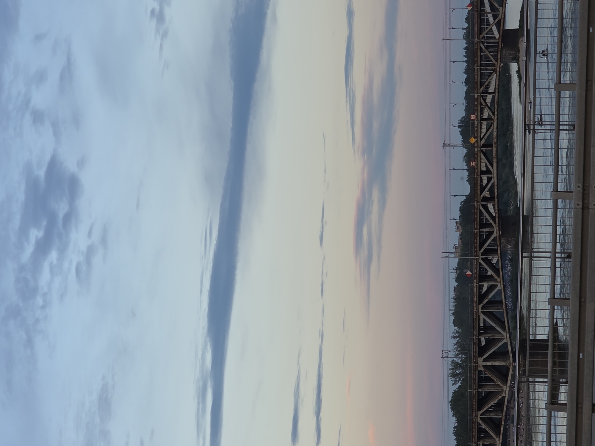 The image captures a view of the Poniatowski Bridge in Warsaw, Poland, spanning the Vistula River. The sky above displays a gradient of colors, transitioning from a pale blue at the top to soft pinks and oranges near the horizon, indicative of either sunrise or sunset. Wispy clouds are scattered across the sky, with some appearing as long, thin bands.
The bridge itself is a prominent steel truss structure, showing signs of age with its weathered appearance. It supports railway tracks and overhead power lines. The river below reflects the colors of the sky and appears calm. Along the riverbanks, a line of trees forms a green backdrop. In the foreground, a metal railing of a pedestrian walkway or another bridge can be seen. There are a few people visible on the riverbank below the bridge, appearing as small figures. Small boats or watercraft are present on the river. The overall impression is a tranquil urban landscape at dusk or dawn.
