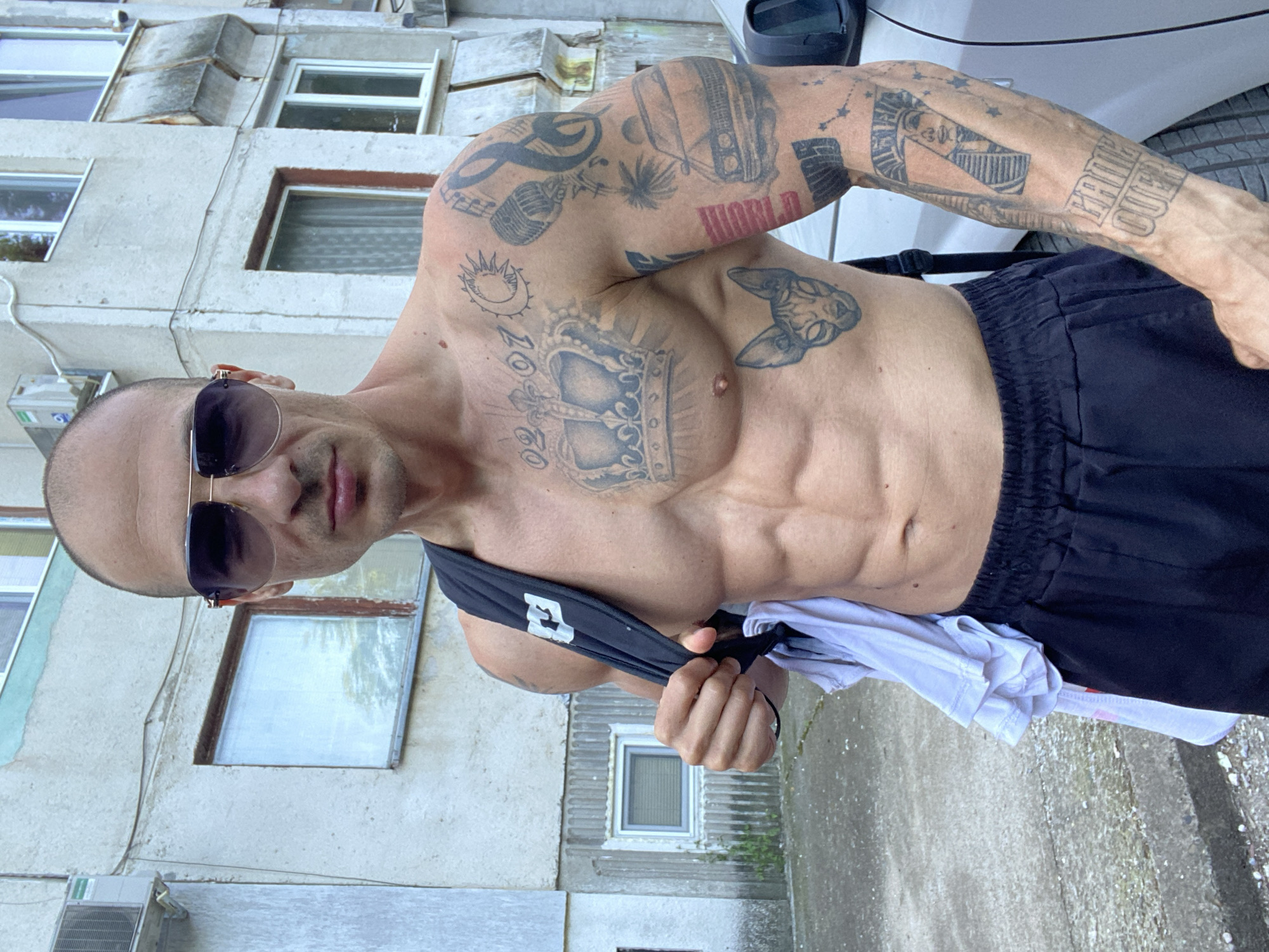 A bald, muscular man with sunglasses and numerous tattoos stands outdoors in front of a dilapidated apartment building in Pleven, Bulgaria. The man is shirtless and wearing black athletic shorts. A black backpack with a white logo is slung over his left shoulder, and a white t-shirt is draped over the backpack strap. The building behind him has peeling paint, exposed concrete, and several windows, some with grime-covered panes. A car is partially visible to the right of the frame. The ground appears to be paved with concrete or asphalt. The lighting suggests it is daytime.