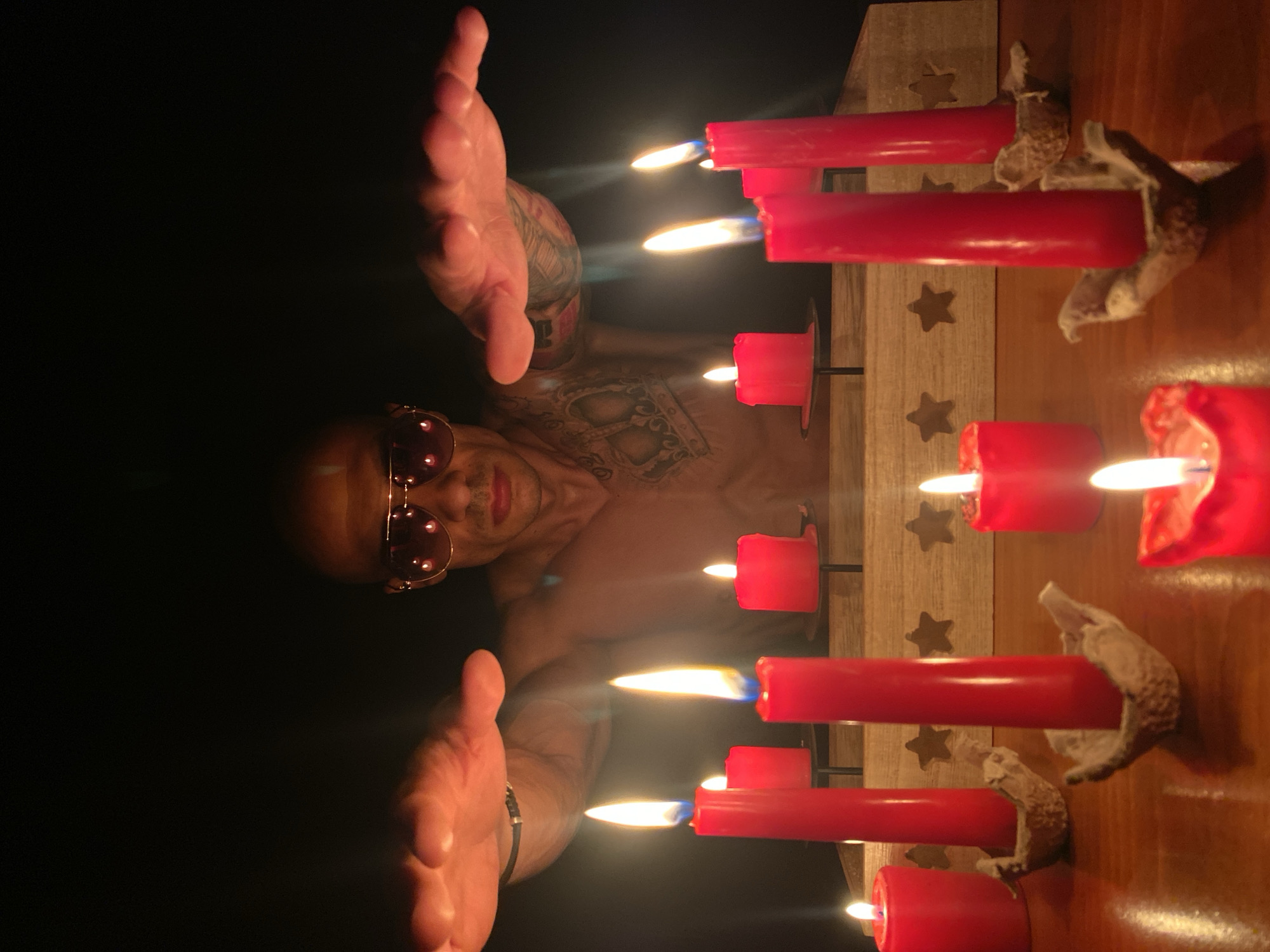 The image depicts a close-up, low-angle shot of a shirtless man with sunglasses and tattoos on his arms and chest, his hands raised over a collection of lit red candles. The man's head is bald, and he is looking directly at the camera. The candles are of various sizes and are arranged on a wooden surface, some in simple holders. The background is dark and out of focus, emphasizing the candles and the man. The scene appears to be indoors, possibly a dimly lit room. There is no indication of a specific location in Pleven, Bulgaria.