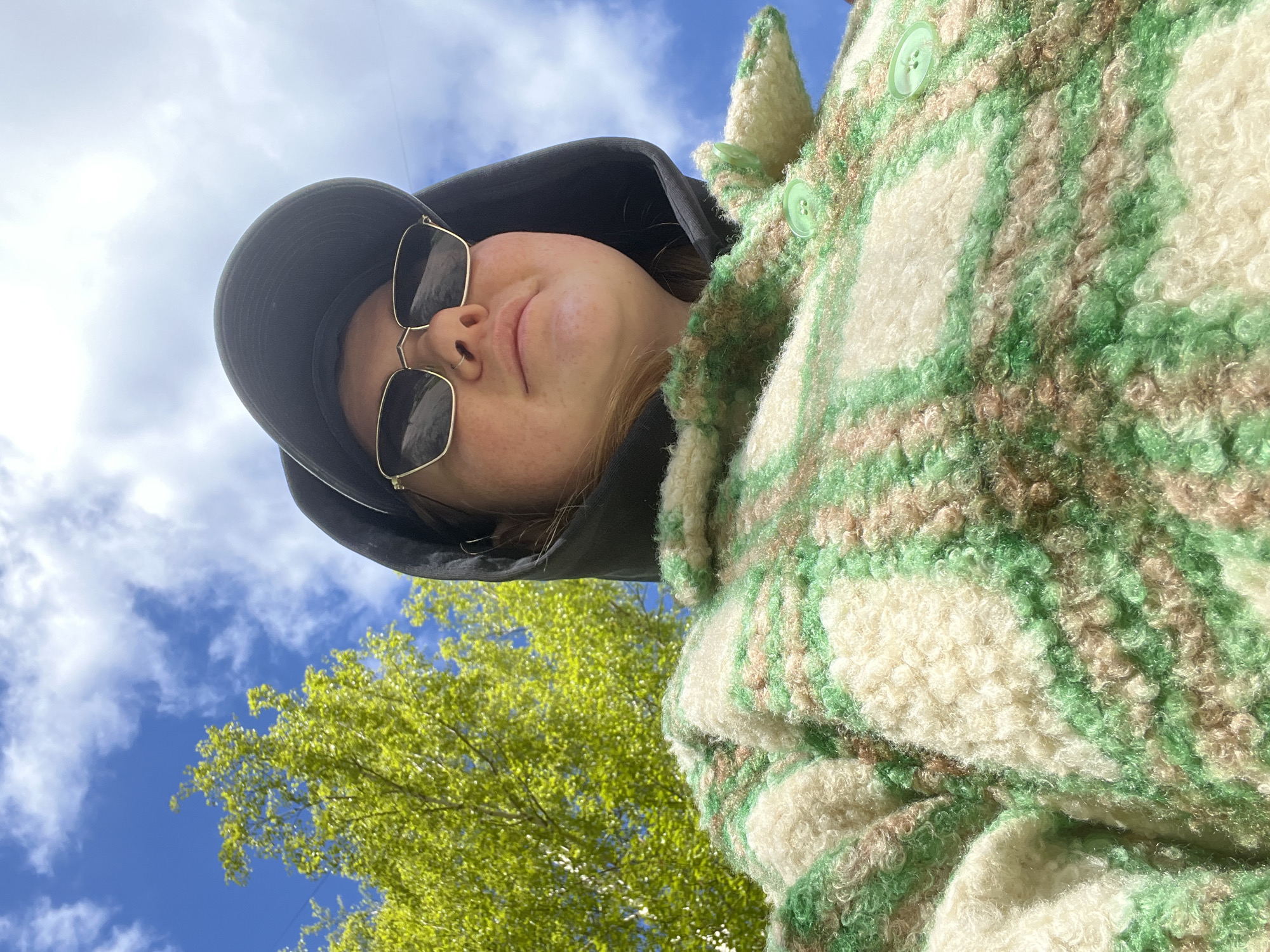 A young woman with fair skin, wearing sunglasses, a black hooded sweatshirt, and a green and tan plaid jacket, is photographed from a low angle. The background shows a blue sky with scattered white clouds, and the green foliage of a tree. The woman is smiling slightly. The location cannot be determined from the image. The picture does not show the city of Chaikovsky, Russia.