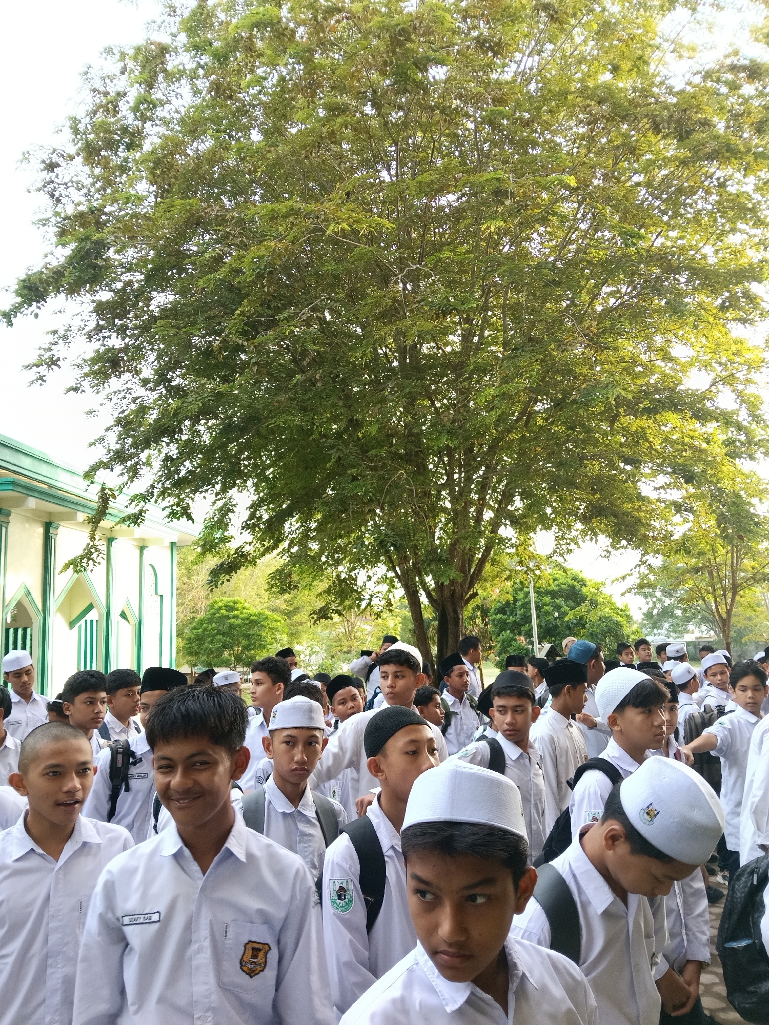 The image shows a large group of male students in white shirts and various head coverings, likely Muslim prayer caps or skullcaps, standing in an outdoor setting. Many have backpacks, suggesting they are students. The setting appears to be a school or religious institution, given the architectural features of a building on the left with green and white trim. A large tree is in the background, occupying a significant portion of the frame. The photo was taken in Lhokseumawe, Indonesia.