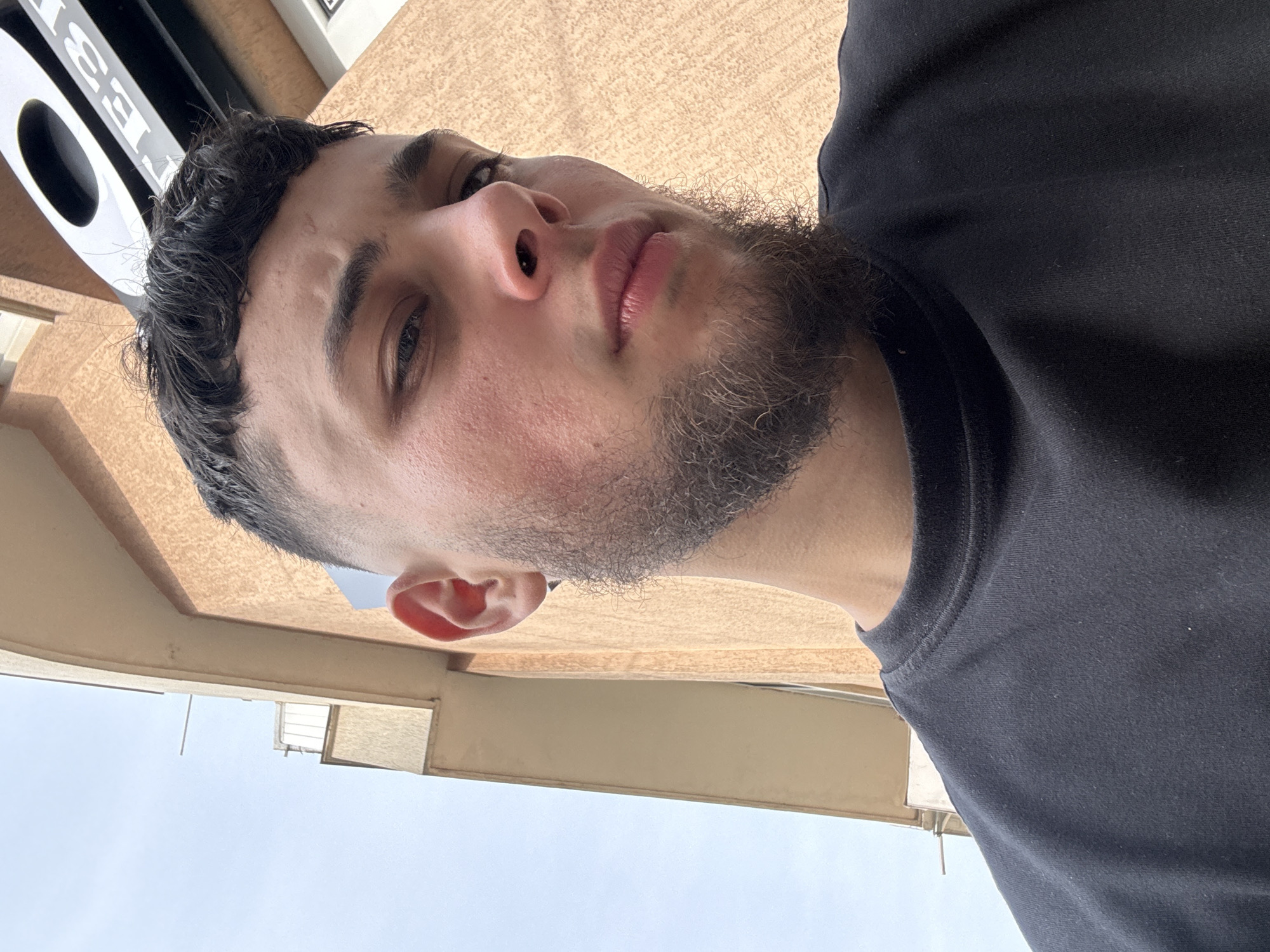 The image is a low-angle selfie of a young man, presented in a portrait orientation but appearing rotated 90 degrees counter-clockwise. The man is captured from the chest up, looking slightly upwards towards the camera with a neutral expression. He appears to be in his late teens or twenties, with short, dark, curly hair and a short, dark, patchy beard. He is wearing a plain black t-shirt. His right eye is visible and dark.
From this low perspective, the background features a light brown, textured overhead structure, possibly an awning or ceiling, positioned above and behind him. Below him, a wide expanse of clear, bright blue sky with no clouds is visible, suggesting a sunny day. Faint architectural outlines of distant buildings can be seen against the sky, indicating an outdoor urban or suburban setting in Varna, Bulgaria.
In the top left of the image (as presented), a white and black object is partially visible. It appears to be a sign or part of a larger structure, displaying the bold, blocky letters "T", "E", "E", "S". The bright, even lighting throughout the scene confirms it is daytime.