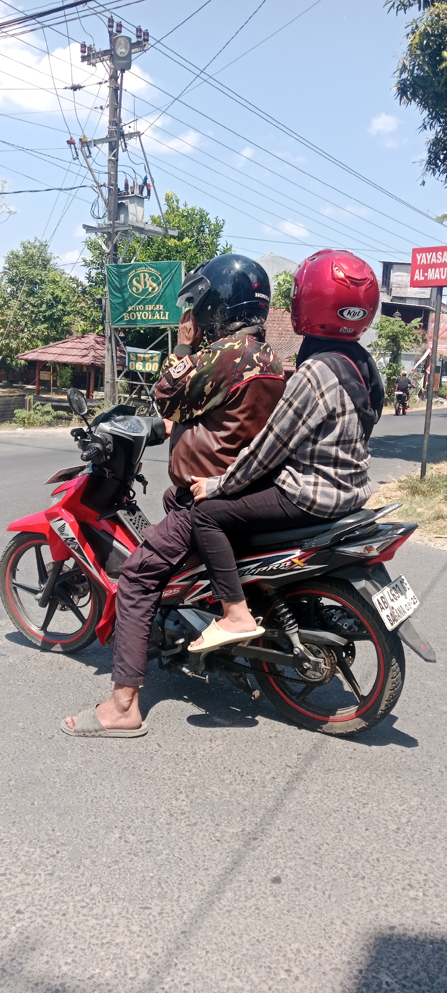 Two people are riding a red motorcycle on a paved road in Maguwoharjo, Indonesia. The person in front is wearing a camouflage jacket and a black helmet, and the person behind is wearing a plaid shirt and a red helmet. They are both wearing casual clothing. The environment appears to be a semi-urban area with trees and buildings visible in the background. There is a utility pole with many wires overhead. A green sign that reads "SOTO SEGER BOYOLALI" is mounted on a pole. A street sign that reads "YAYASAN AL-MA'ARIF" is also visible. The sun is shining, casting shadows on the road. The motorcycle's license plate reads "AB 4683 RE" and "08-29".