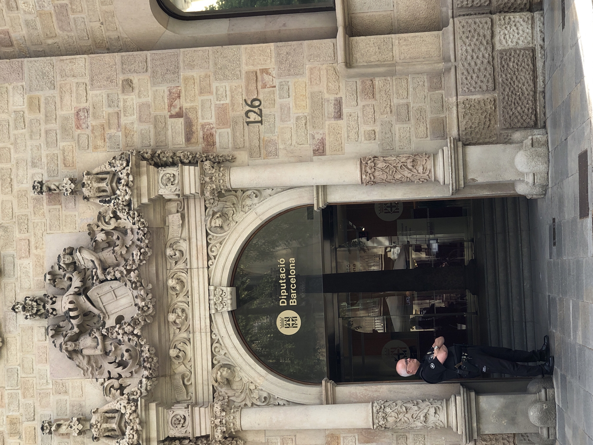A male security guard, wearing a black uniform with a badge on his left chest and a gun holster on his right hip, stands with his arms crossed in front of an arched entrance. The entrance is a glass door with the "Diputació Barcelona" logo. The building is made of stone blocks and features elaborate carvings above the entrance, including figures, a coat of arms, and floral motifs. The number "126" is visible on the stone wall to the right of the entrance. The ground in front of the building is paved with stone.