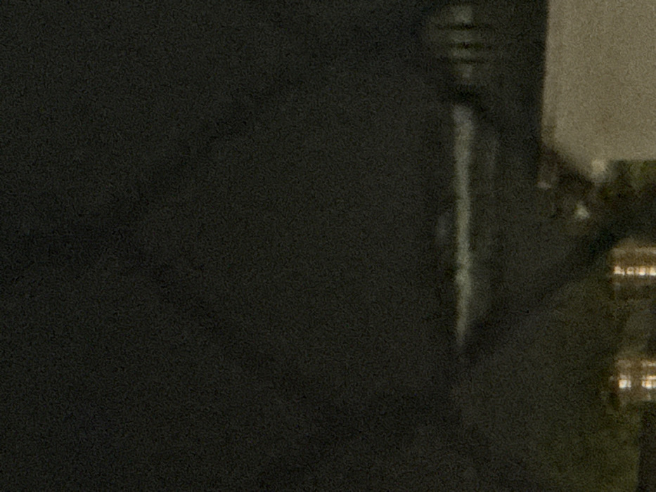 The image depicts a dark, grainy, and blurred scene at night in Ngũ Hành Sơn Ward, Vietnam. A prominent horizontal band of white and light grey blur extends across the mid-ground, consistent with breaking waves on a body of water, likely the ocean. Above this, the sky is uniformly dark. In the lower left foreground, several indistinct, warm-toned (orange/yellow) lights are visible, indicating artificial illumination from structures or streetlights. To the lower right, a portion of a light-colored, solid structure, possibly a building or wall, is partially visible. Faint, dark vertical blurs to the right of the wave line may represent distant structures or vegetation. No individuals, specific actions, or interactions are discernible.