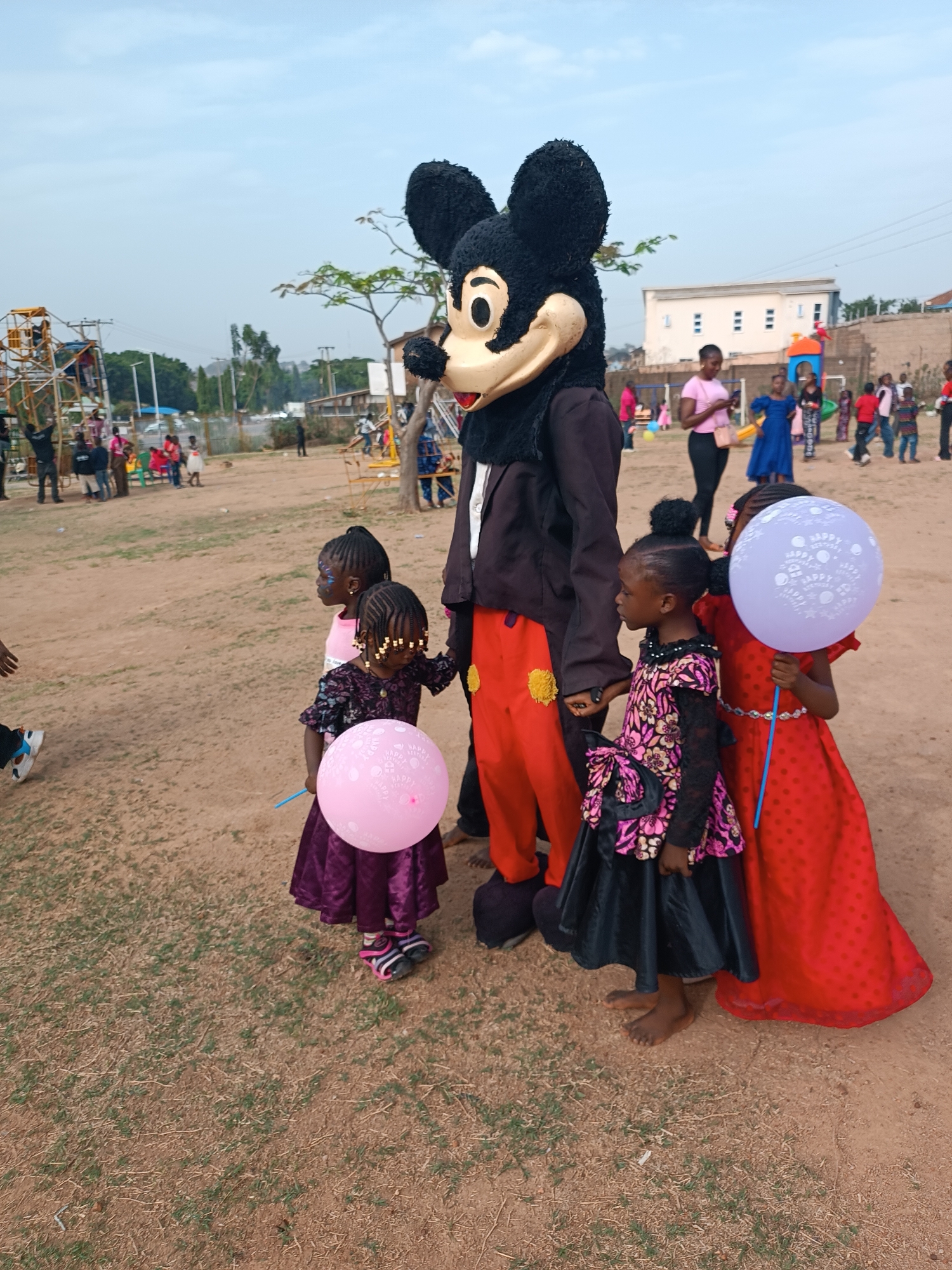 In an outdoor setting in Jos, Nigeria, a costumed character resembling Mickey Mouse stands surrounded by three young girls. The character is wearing a black jacket, orange pants, and oversized black shoes. The first girl, in a purple dress, holds a pink balloon. Next to her is another girl with face paint and braids with beads, also wearing pink. The third girl wears a dress with black lace sleeves and a pink floral pattern, followed by a girl in a red dress holding a blue balloon with white prints.
In the background, there are other people, a building, trees, and an amusement park-style ride. The ground is a mix of dirt and grass.