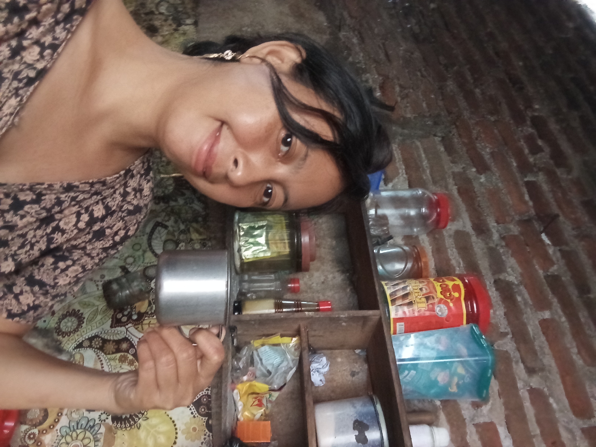 The image shows a woman holding a silver metal object, possibly a grinder, in front of a wooden shelving unit. The unit contains various items including glass jars, plastic containers, and small miscellaneous objects. The woman is positioned at an angle, with her face visible and smiling. The background is a brick wall, and the setting appears to be indoors. The image includes a patterned cloth underneath the shelving unit. There's no definitive information to indicate the location as Baron, Indonesia, from the image alone.