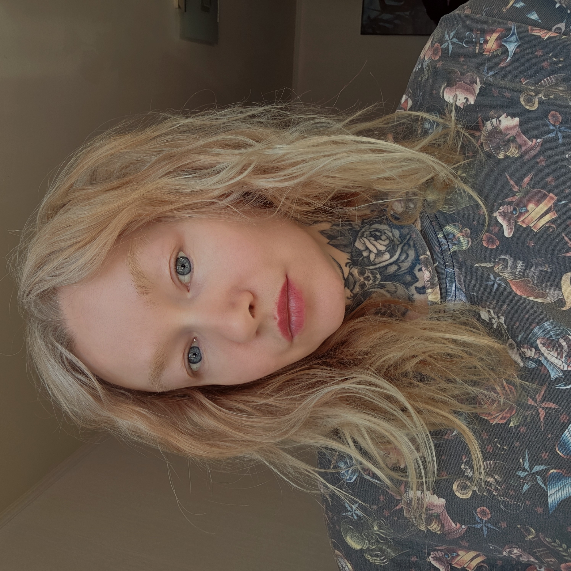 A young woman with light blonde hair, blue eyes, and fair skin poses indoors. She has a neck tattoo featuring a rose and skulls. She's wearing a dark patterned top. The background is a neutral wall color. The location cannot be determined as Saint Petersburg, Russia.