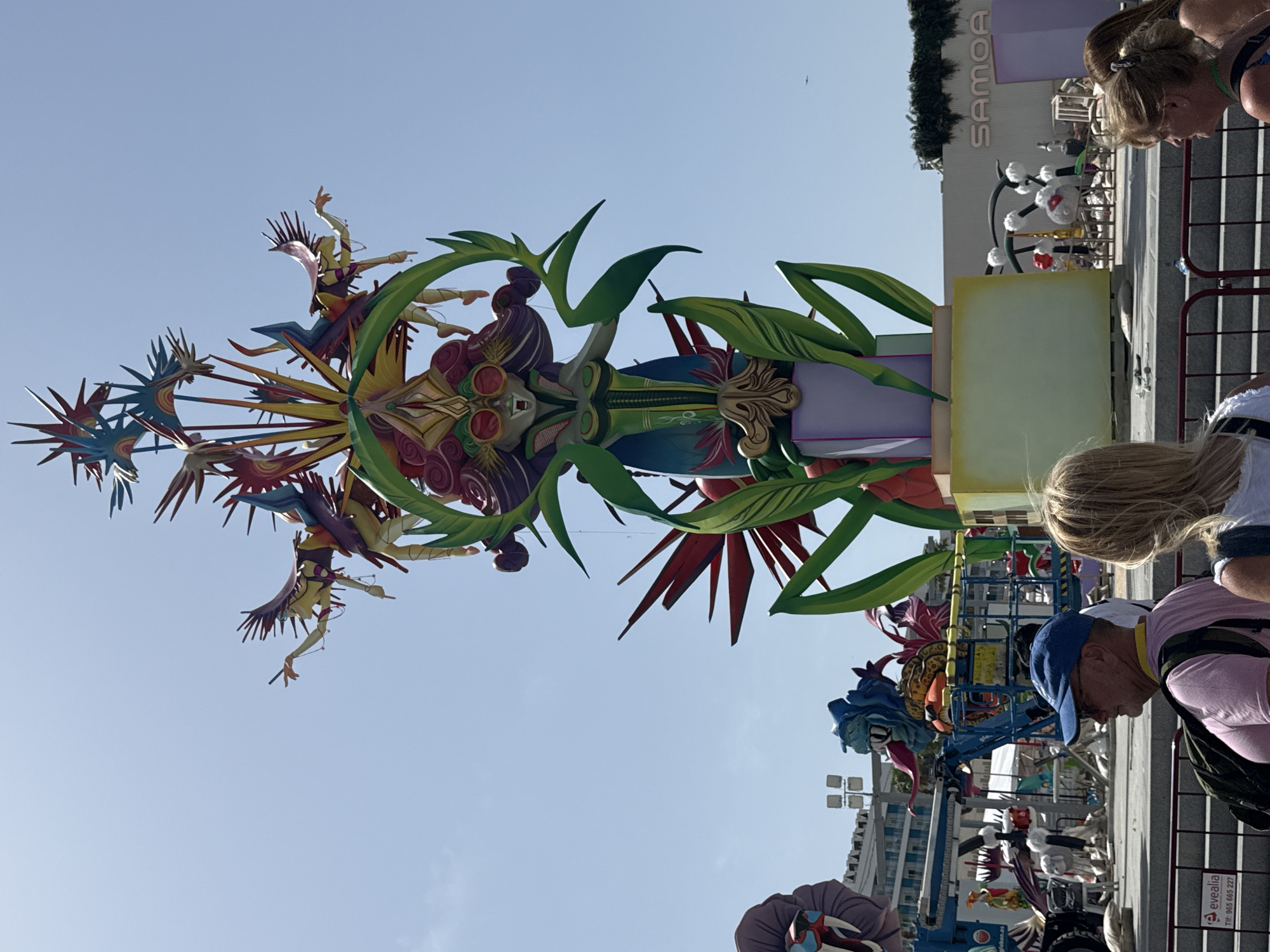 A large, ornate sculpture stands tall against a clear blue sky. The sculpture depicts a female figure with striking makeup, wearing sunglasses, and adorned with vibrant, colorful wings and leafy green appendages. The figure is situated on a platform, which is part of a larger, elaborate structure. In the background, various other colorful sculptures and structures are visible, some appearing to be under construction or being assembled. There are also several people in the foreground, some looking at the sculptures, and one man wearing a blue cap and pink shirt. The overall environment appears to be an outdoor public space, possibly a festival or exhibition. The sculpture is the main subject, with intricate details and a fantastical, artistic design.