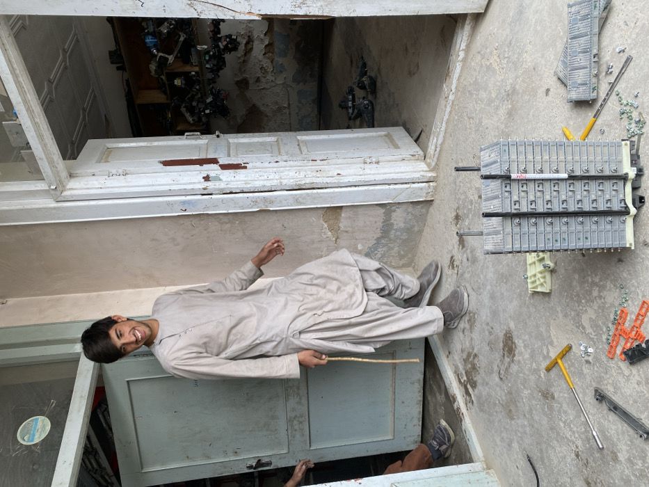 The image depicts an informal workshop or storage area in Kandahar, Afghanistan. In the central foreground, a multi-cell hybrid vehicle battery pack, likely from a Toyota Prius, is partially disassembled on a concrete floor. It consists of approximately twenty grey modules stacked vertically, secured by two black rods. Scattered around it are various tools and components, including a yellow T-handle wrench, a silver extension, several small bolts and nuts, and orange plastic battery cell spacers. Another section of grey battery modules is visible to the right of the main stack.
A young adult male, dressed in a light-colored shalwar kameez and grey slip-on shoes, stands in the left portion of the image, partially within an open, light blue-green wooden doorway. He is smiling and looking towards the left, appearing in motion with his right leg slightly lifted. He holds a slender, light-colored stick-like object in his left hand. Part of another individual's leg and foot is visible inside the left doorway.
To the right, another white wooden doorway, showing significant paint chipping and some exposed reddish-brown underlayer, stands open. This doorway leads into a cluttered, dilapidated room with peeling paint and crumbling plaster walls. Inside, numerous dark, unidentifiable electronic or mechanical components are stacked on shelves and scattered on the floor. The ceiling of this interior room features a patterned design. The wall between the two doorways is a light, unpainted surface. A small, circular, light blue sticker is affixed to the upper left door frame/wall.