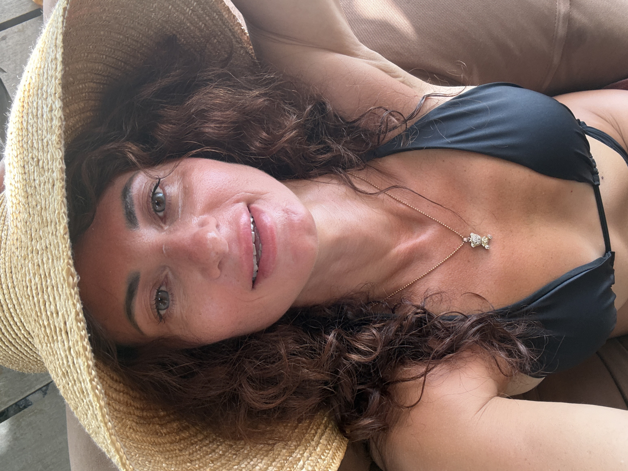 A woman wearing a wide-brimmed straw hat and a black bikini top is shown in a close-up, selfie-style portrait. Her curly, dark brown hair frames her face, and she is wearing a delicate gold necklace with a small, jeweled pendant. The background appears to be a neutral-colored surface, possibly furniture. The lighting suggests an outdoor or well-lit indoor setting. The image was taken in Baan Tai, Thailand.