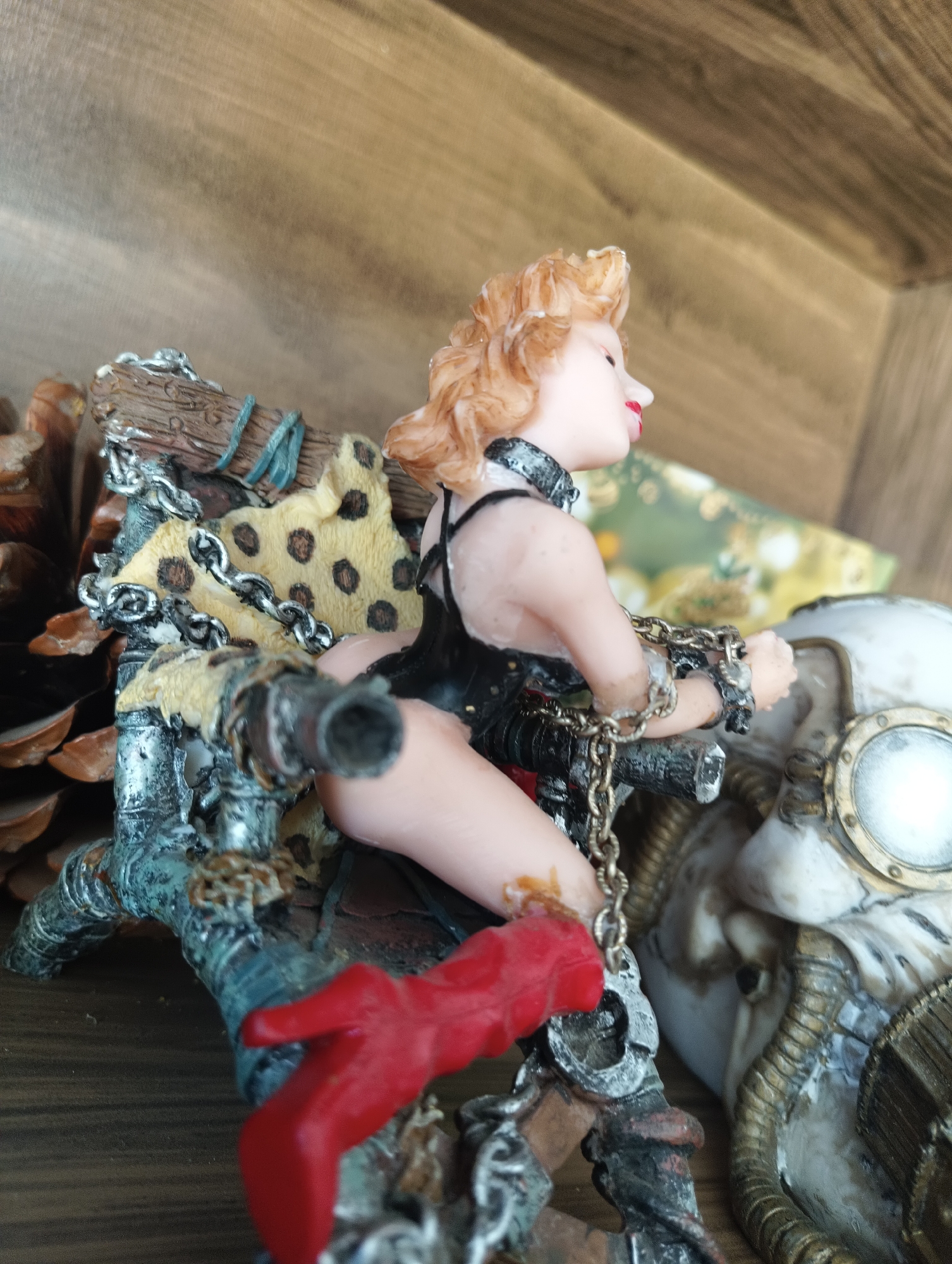 This is a close-up, medium-angle, eye-level shot of a figurine. The figurine depicts a stylized, blonde woman with reddish-pink lips and rosy cheeks, wearing a black, low-cut outfit and a studded collar. She is chained at the wrists with a metal chain that is attached to a rustic, wooden structure. This structure is adorned with blue twine, a piece of leopard-print fabric, and metallic, grey-colored elements that resemble pipes or metal plating. A red, high-heeled shoe is visible at the bottom left of the frame, attached to the wooden structure. To the right of the figurine, a steampunk-inspired object with a large, round, metallic-rimmed lens is partially visible. The background consists of a wooden surface, possibly shelves or a wall, with a slight tilt to the right. The lighting suggests an indoor setting with soft illumination. The overall aesthetic is dark and industrial with elements of fantasy or fetish art. No visible text is present in the image. The location context of Karnobat, Bulgaria, does not provide further visual cues within the image itself.