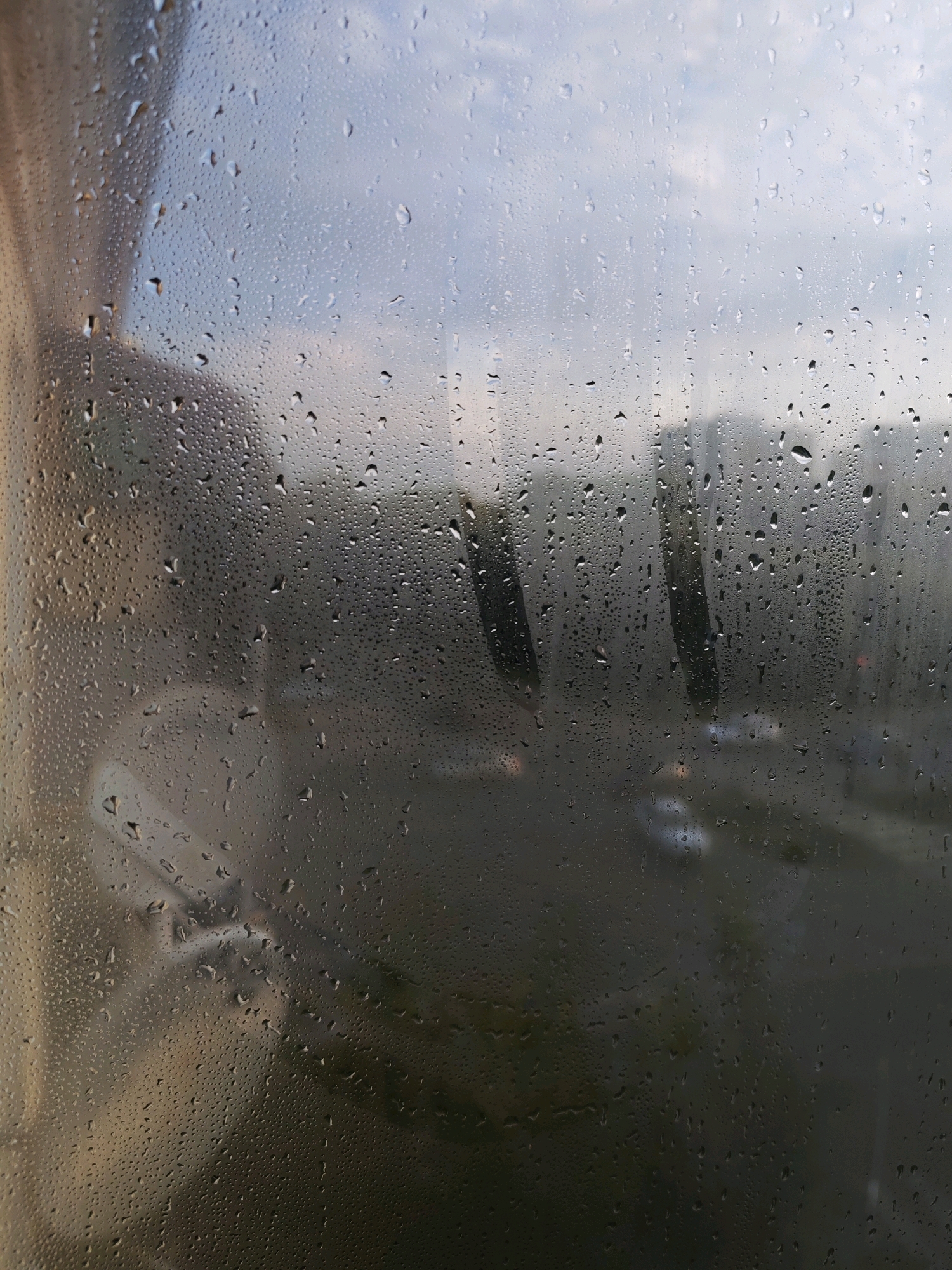 The image depicts a view through a window covered in condensation and water droplets, looking out onto a city street in Izhevsk, Russia. The exterior scene is blurred and out of focus due to the water on the glass. Buildings, some with visible windows, are discernible in the background, along with indications of vehicles on what appears to be a road. The sky is overcast, suggesting a cloudy or rainy day. In the foreground, on the windowpane itself, there are streaks and patterns formed by the water droplets. The overall impression is one of looking out from an indoor space onto a muted, urban landscape on a damp day.