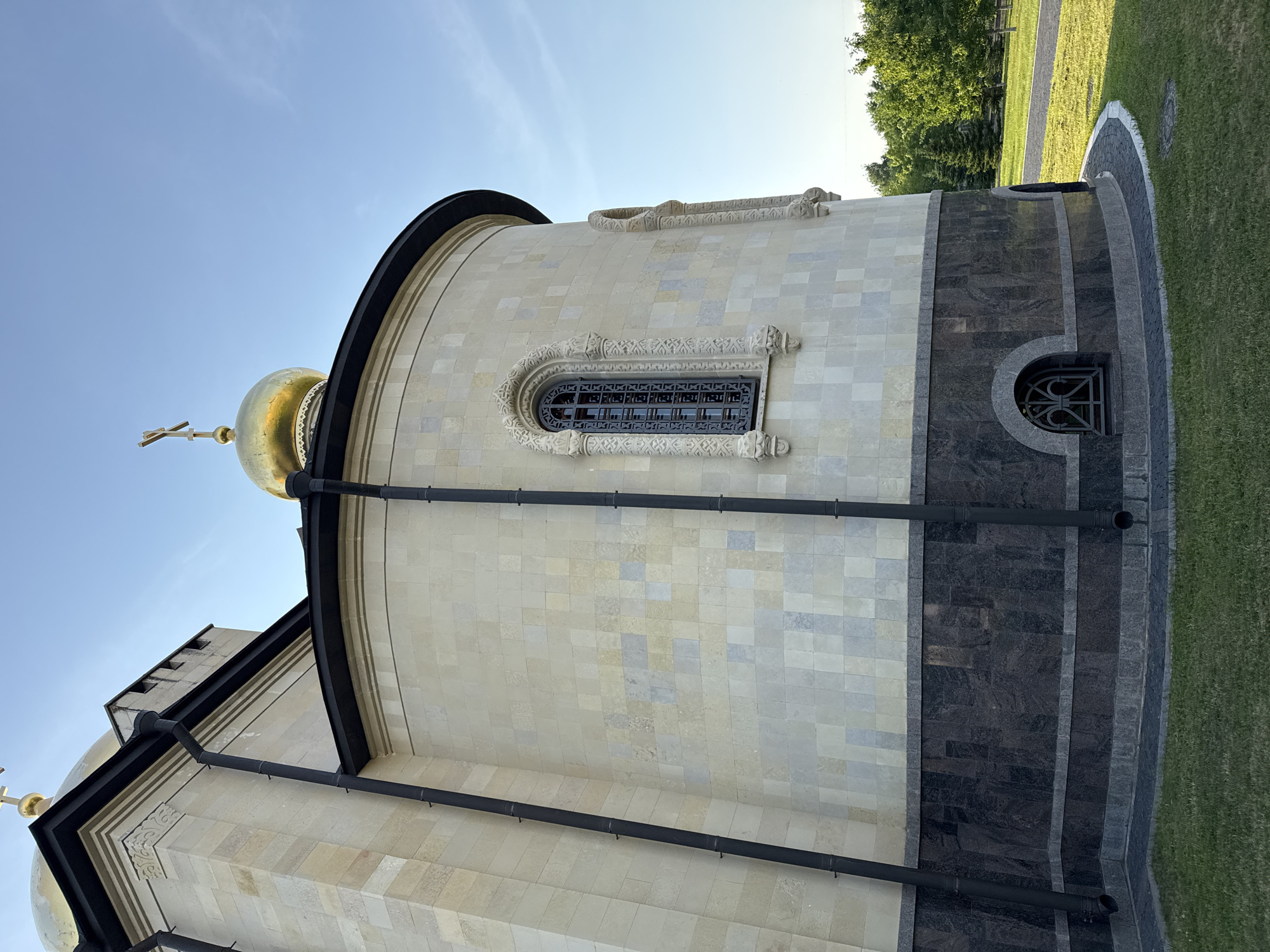 The image shows the exterior of a building, possibly a church, with a golden dome topped with a cross, visible against a clear blue sky. The building's facade is constructed of light-colored stone tiles arranged in a mosaic pattern. A dark-colored band runs around the lower portion of the building, above which is a section of dark granite with a lighter granite trim. Two black downspouts run vertically down the side of the building. There is a small arched window with a decorative metal grate on the lower part of the building. A paved path and manicured grass are visible in the foreground and to the right, with a line of trees in the background. This is the Church of the Assumption of the Blessed Virgin Mary in Kirishi, Russia.