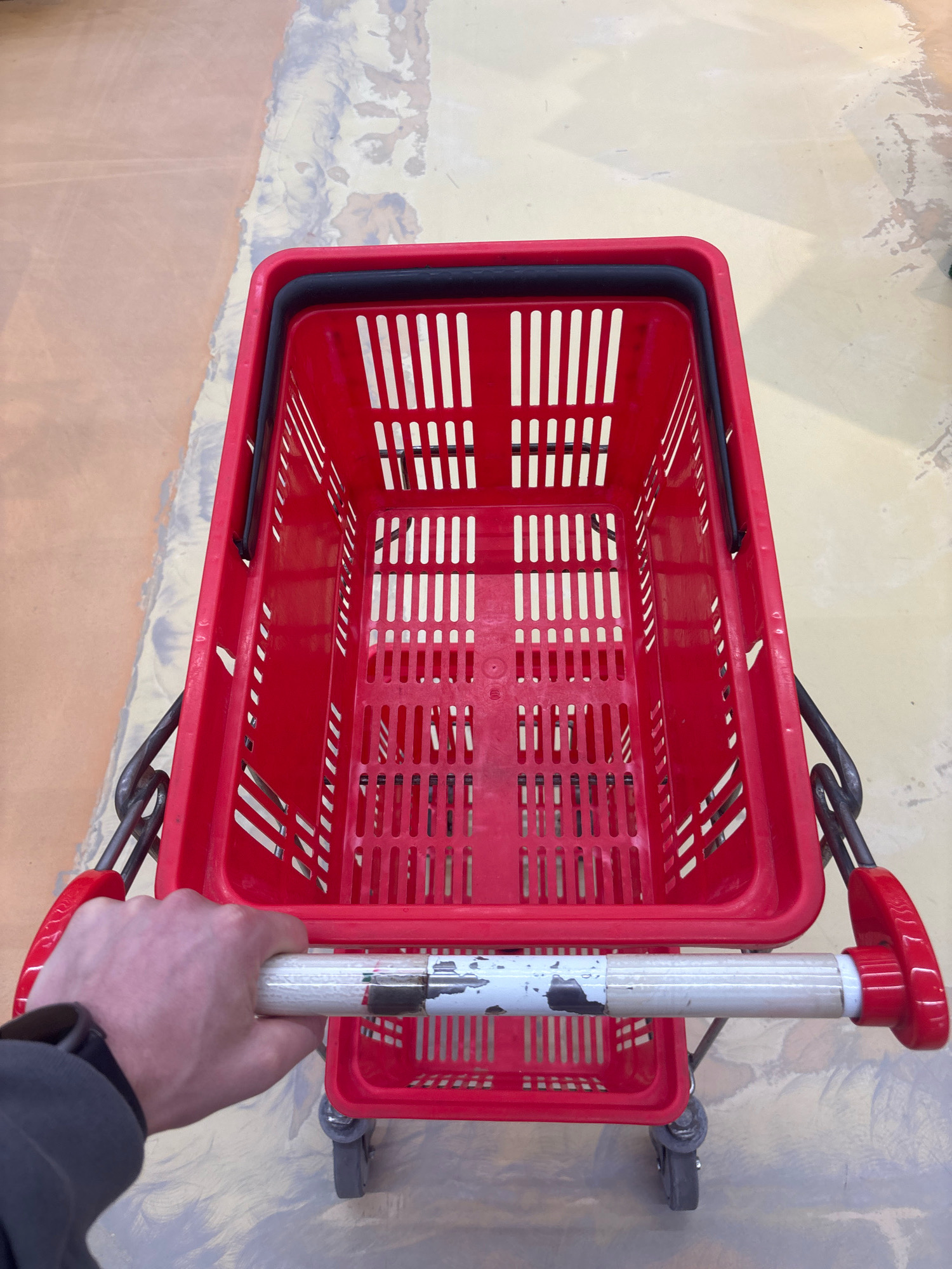An overhead, close-up shot shows a red plastic shopping cart from the perspective of someone holding the handle. The cart is empty and sits on a floor that appears to be concrete with patches of discoloration and wear. The cart has a black rim around the top of the basket and two handles on the sides. The handle being held is white with some wear and tear, showing patches of darker material underneath. The background shows the transition from a lighter-colored floor to a darker, more orange-toned floor. No people are visible other than the hand holding the cart.