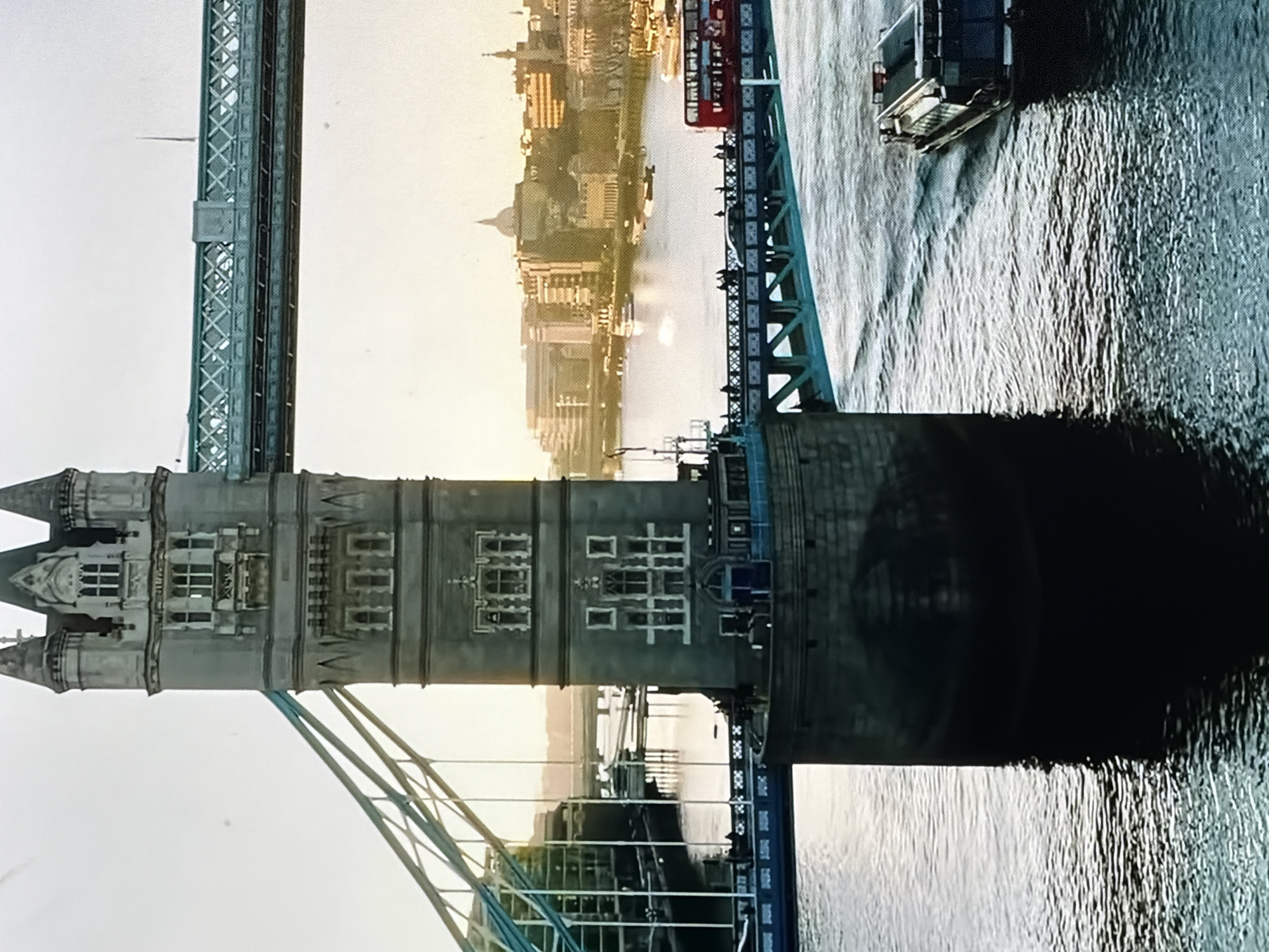 The image presents a detailed view of a large, stone-built bridge with distinctive Gothic-revival twin towers. The bridge spans a wide body of water, with the substantial base of the foreground tower prominently reflected. On the bridge deck, a red double-decker bus travels alongside several pedestrians. A modern boat navigates the water away from the bridge, creating a visible wake. In the background, a cityscape extends along the river, featuring numerous buildings and a notable dome-topped structure. The sky above is bright with a soft, warm light, indicating either early morning or late afternoon. This scene is located in the city of Maidstone, United Kingdom.