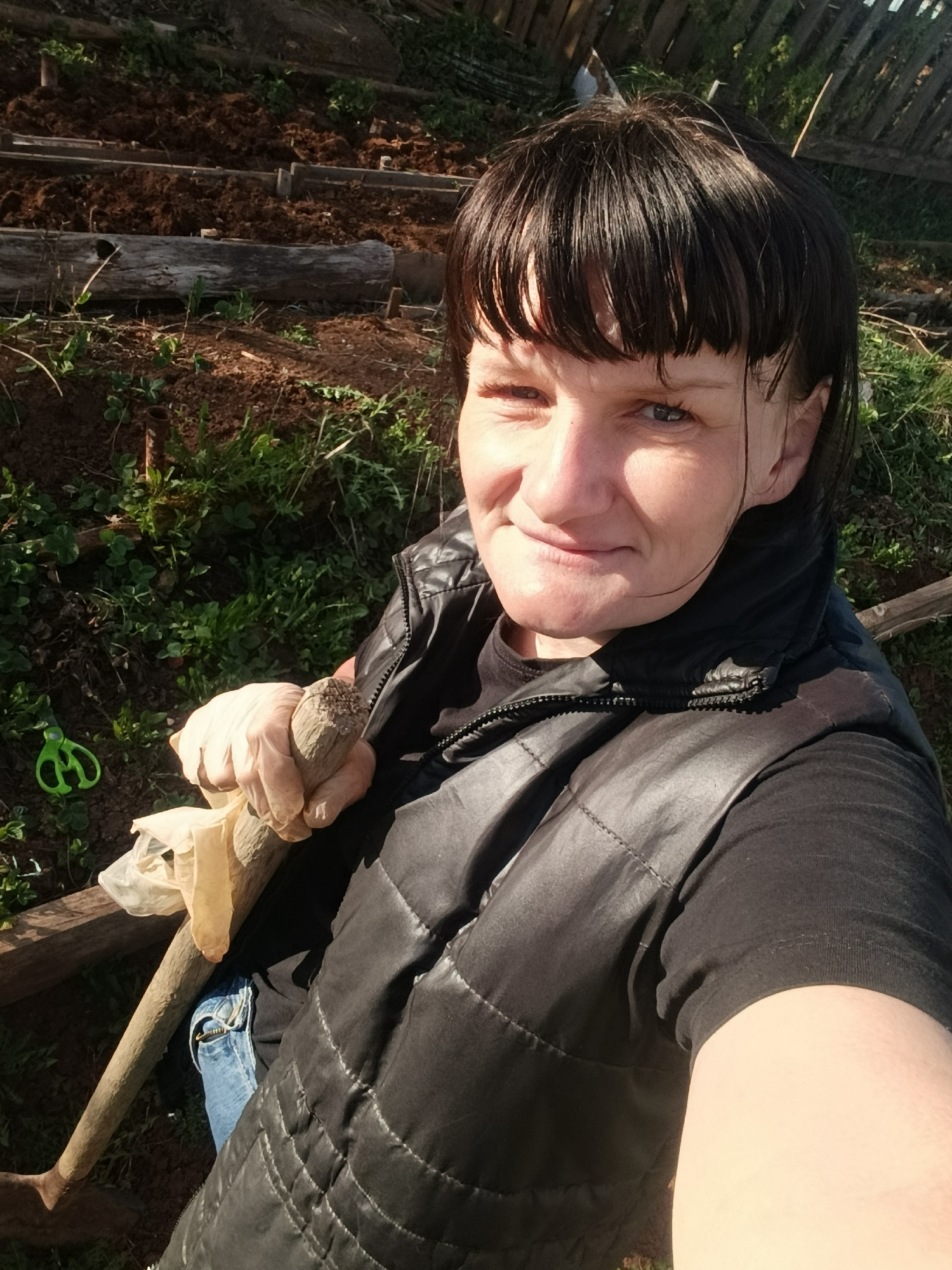 A woman is outdoors in a garden. She is fair-skinned with short, dark hair and blue eyes. She is wearing a black vest, a black T-shirt, and blue jeans. She is holding a wooden-handled shovel in her gloved hand. Behind her are raised garden beds with tilled soil and patches of green vegetation. A green pair of scissors is visible on the ground near the shovel. A wooden fence is partially visible in the background. The photograph appears to be a selfie taken by the woman.
I am unable to determine the location of this photo to Фоки, Russia.