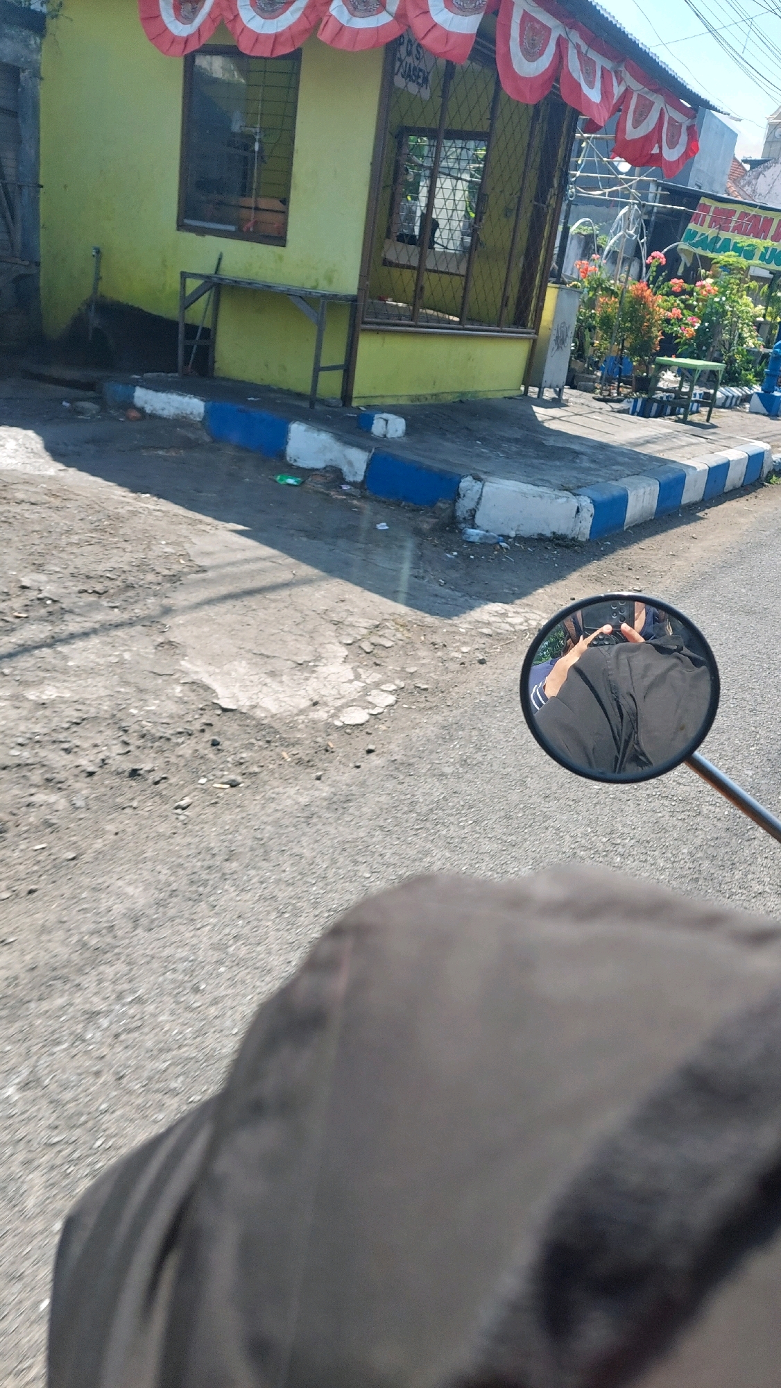 The image depicts a street scene in Sidoarjo, Indonesia, focused on a small yellow building with red and white decorative banners hanging from the roof. The building has windows with metal grates and a small metal table outside. In front of the building is a concrete sidewalk with blue and white painted borders. The road in the foreground is asphalt and appears to be in disrepair. A motorcycle's side mirror is visible, reflecting a person wearing a black garment, presumably the rider. Plants and flowers are visible in pots and in the background, along with other buildings and structures. The overall impression is of a roadside establishment in a semi-urban or rural area.