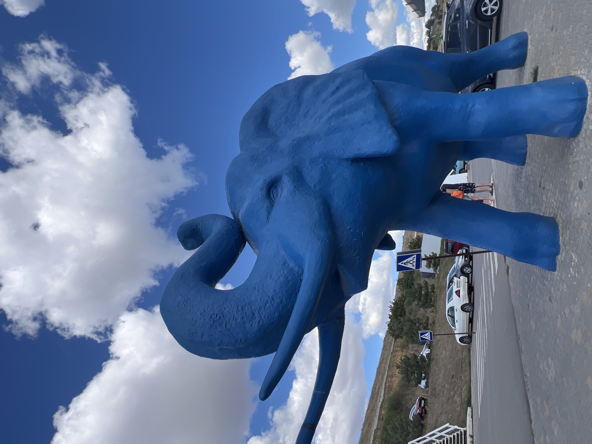 A low-angle, full-shot captures a large, vibrant blue elephant sculpture in the foreground. The sculpture is positioned outdoors, likely in a public space, with a bright blue sky dotted with fluffy white clouds above. The elephant is facing to the left of the frame, with its trunk raised and curved upwards. Its tusks are visible, extending forward. In the background, a road with a pedestrian crossing is visible, with several cars parked nearby. A slight incline with greenery and some trees forms the backdrop behind the road. Two people are standing near the elephant's legs. The scene appears to be in Fontanka, Ukraine, an area known for its seaside attractions.