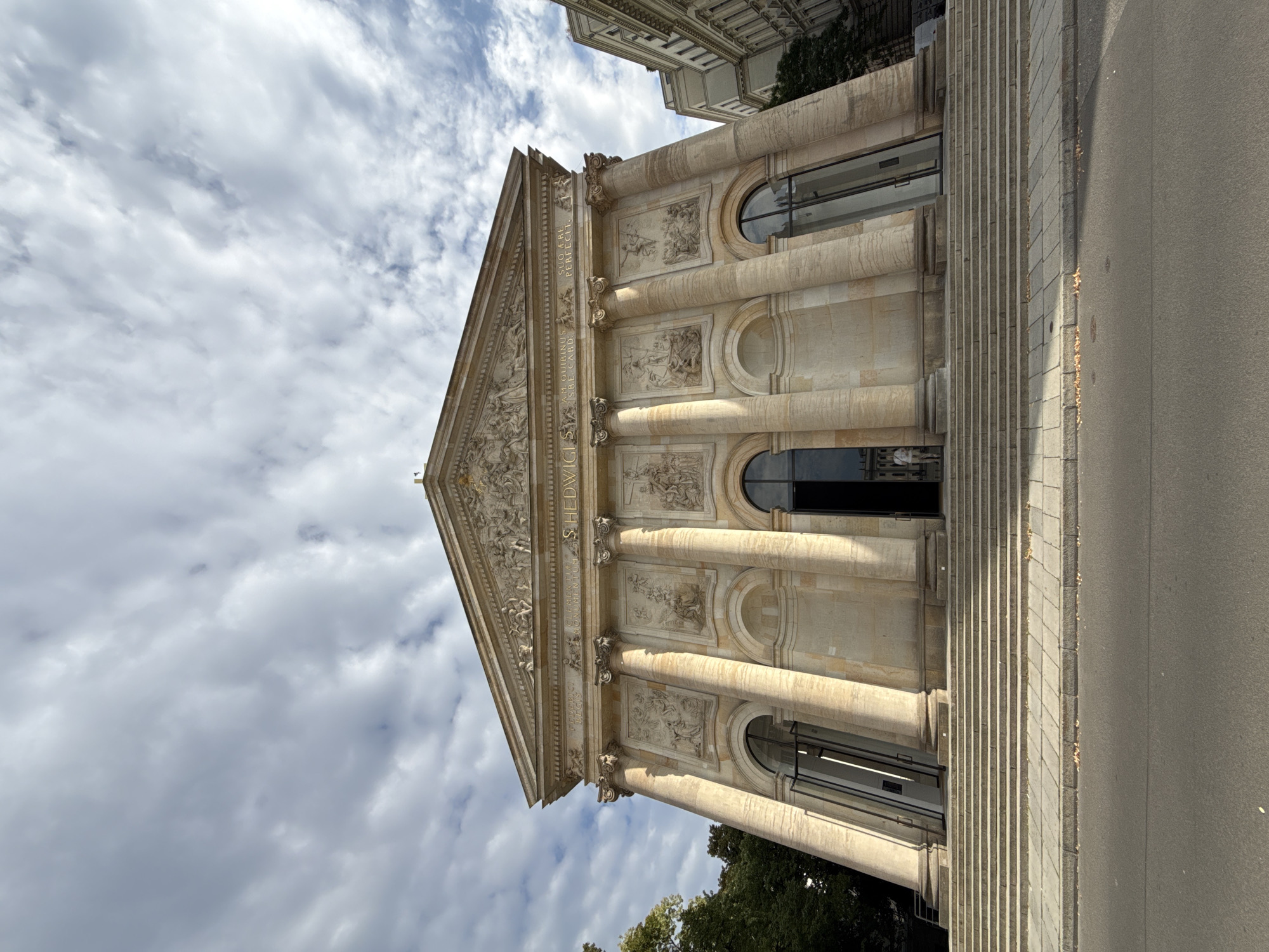 The image shows a low-angle, wide shot of the front facade of the Altes Museum in Berlin, Germany. The neoclassical building features a grand staircase leading up to the entrance, flanked by large columns and ornate friezes. The sky above is partly cloudy, with patches of blue visible. A street is in the foreground, and to the left, trees are visible. A few people can be seen standing near the entrance on the steps. The sun casts shadows on the building, highlighting its architectural details. The overall scene is one of historical grandeur and public space.
